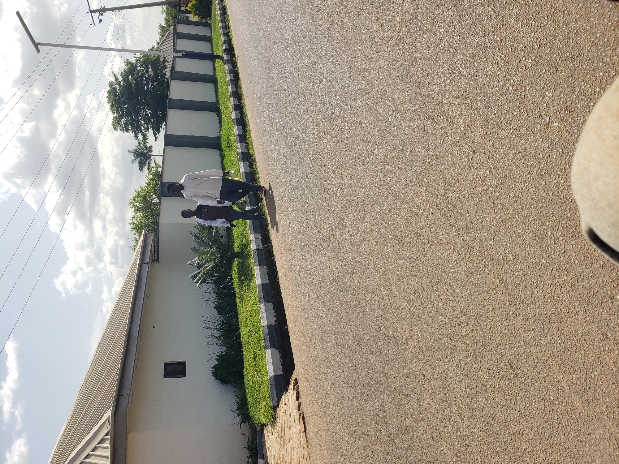 Two young men are walking on a paved road in Gura Top, Nigeria. The man on the left is wearing a white shirt with a pattern and dark pants. He is holding a plastic bag in his left hand. The man on the right is wearing a white long-sleeved shirt and dark pants. He is also holding a plastic bag in his left hand. They are walking on the right side of the road. In the background, there is a white wall with a dark grey trim, and a section of a building with a grey metal roof. There is also lush green grass and some trees. The sky is partly cloudy. The road is paved and appears to be in good condition. There are power lines running across the sky. A lamppost is visible on the right side of the image.
