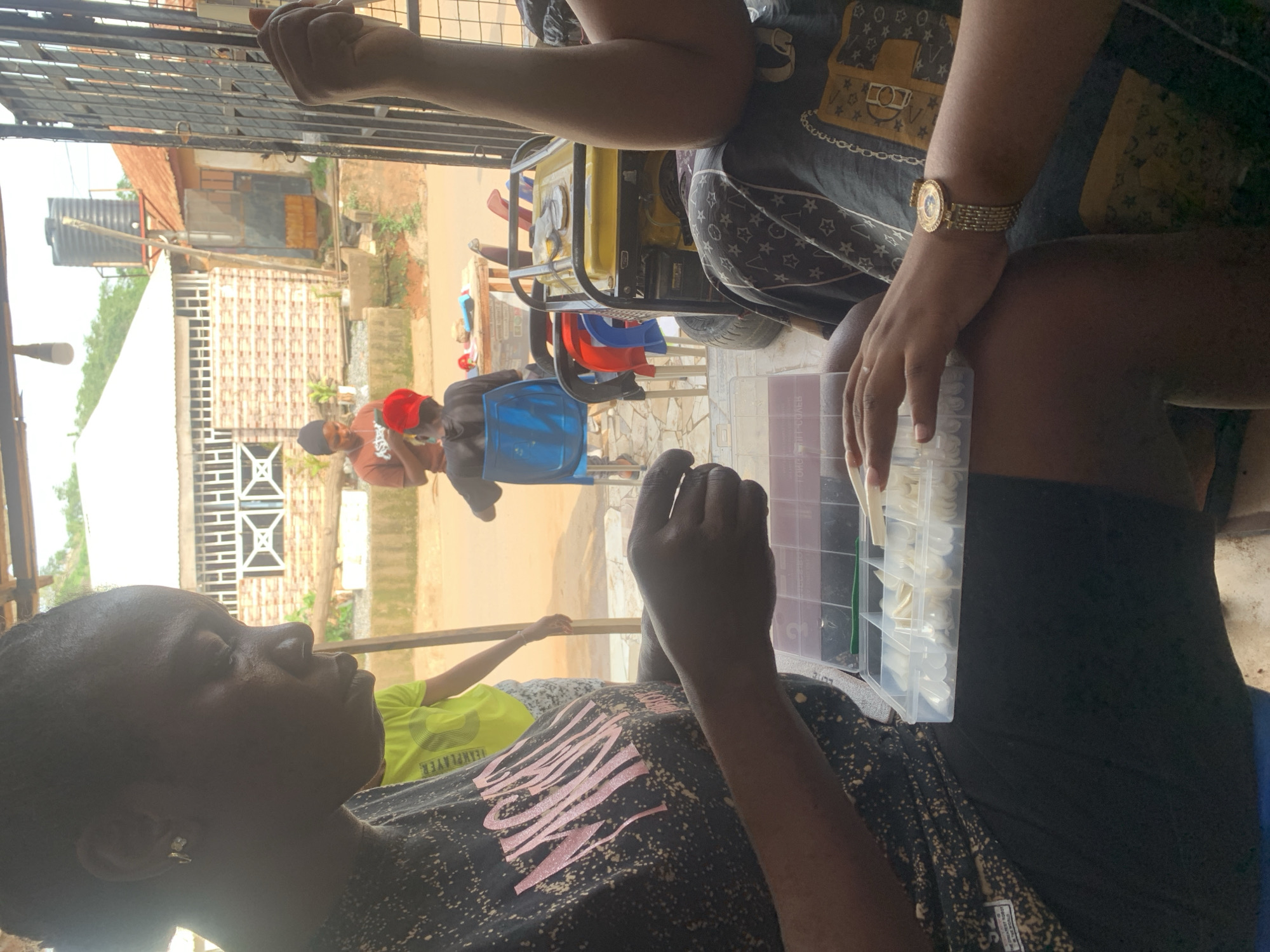 A woman is seated indoors with a container of artificial nails on her lap. Another person sits adjacent to her wearing a gold watch. Outside, two men are seated on blue plastic chairs, and other houses and buildings can be seen. The scene appears to be taking place in Abuja, Nigeria.