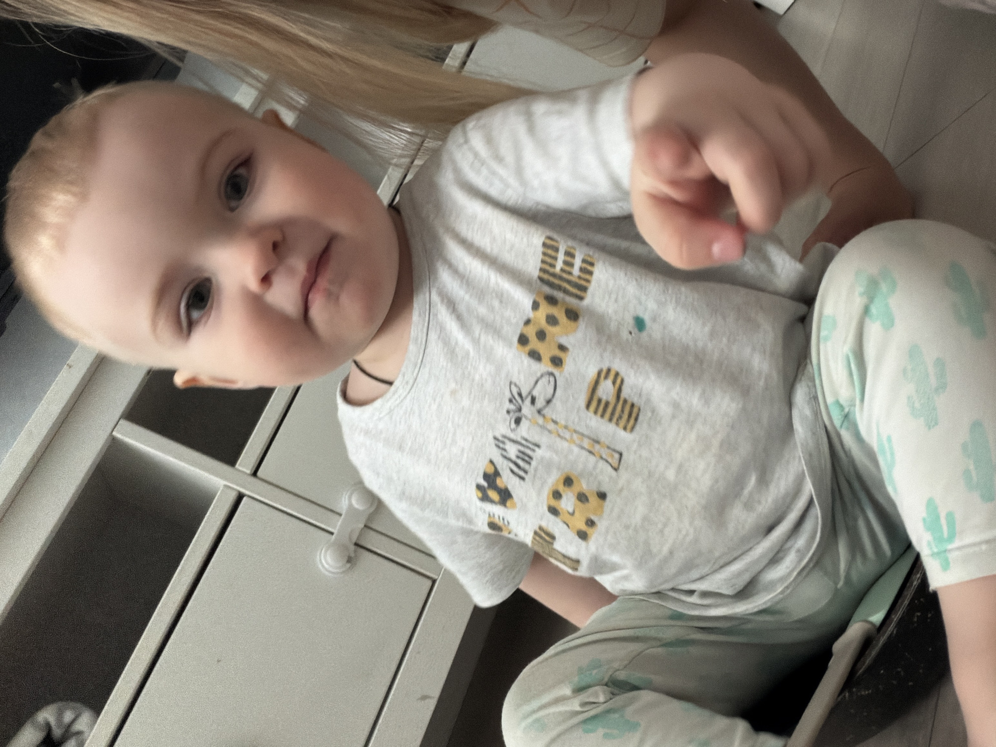 A toddler with fair skin, light blonde hair, and wearing a light gray t-shirt with animal print letters and a giraffe design is seated on the floor. The child is pointing with their right hand towards the viewer. They are also wearing light blue and white cactus-printed leggings. To the left, a white cabinet with drawers is visible. The top of an adult's head and blonde hair appear in the top part of the frame. The location is not identifiable as the city of Votkinsk, Russia, based on the provided image.