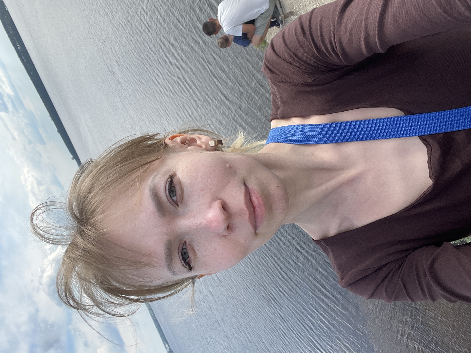 A selfie-style portrait shows a young woman in the foreground, with a vast body of water and distant land in the background. The woman has light-colored hair, which is styled loosely around her face, and light eyes. She wears a dark brown long-sleeved top and a prominent blue strap, possibly from a bag, crosses her shoulder. A small gold-colored earring is visible in her right ear. She looks directly at the camera with a neutral to slightly smiling expression.
In the mid-ground, near the edge of the water, two men and a small child are visible. One man wears a white t-shirt and grey shorts, kneeling or crouching, while the child in a blue t-shirt is close to him. Another man is partially obscured. They appear to be engaged in a quiet activity near the water.
The setting features a large expanse of water, likely a lake or wide river, with subtle ripples on its surface. A strip of land, covered with trees or vegetation, is visible on the distant horizon, separating the water from the sky. The sky above is cloudy, suggesting an overcast or partly cloudy day. The time of day appears to be daytime, with diffused light. The location is specified as Пятый километр, Russia. There is no visible text in the image.