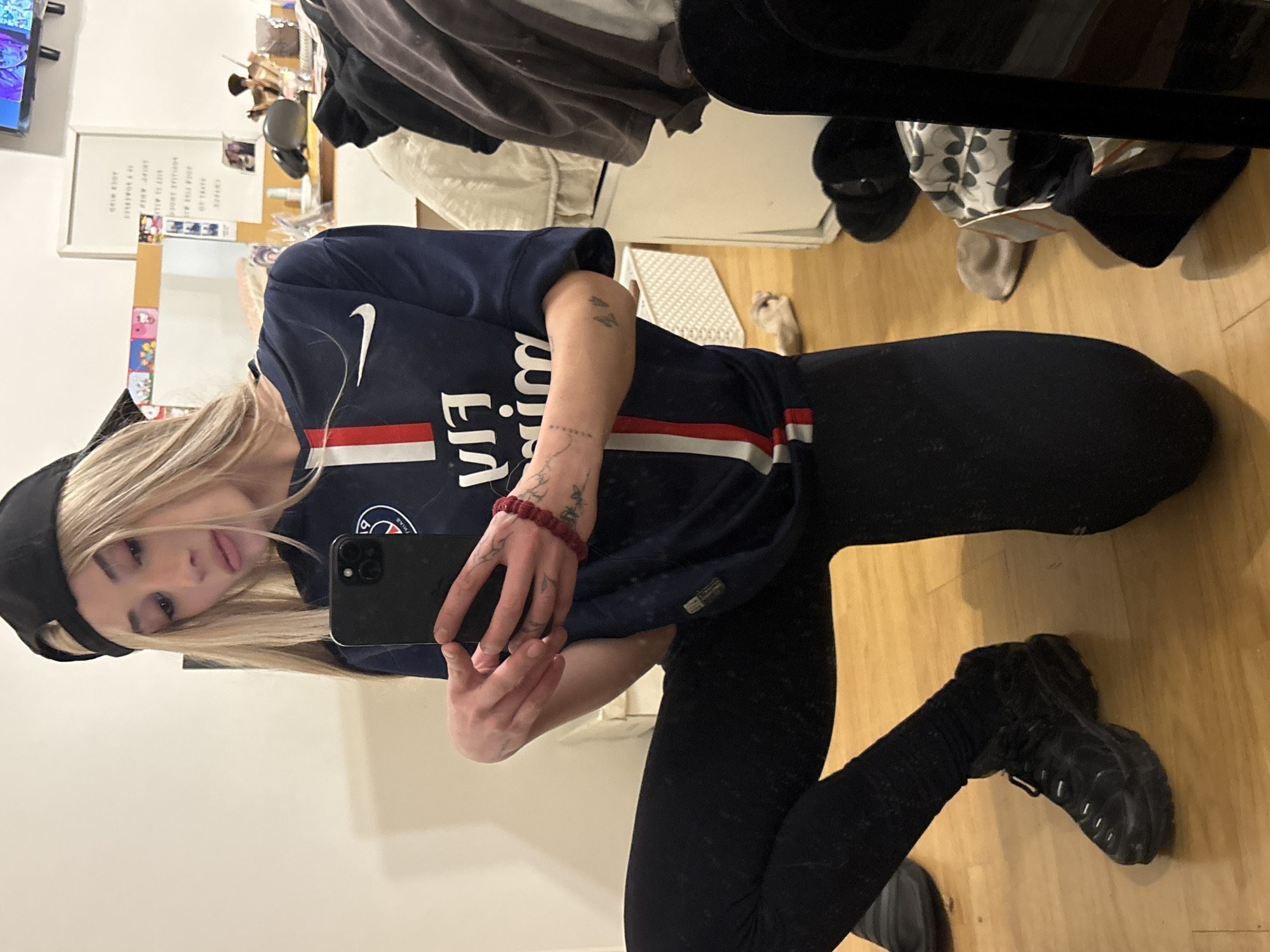 A person is kneeling and holding a smartphone to take a selfie. They are wearing a black baseball cap, a dark blue Paris Saint-Germain soccer jersey, black leggings, and black boots. They have blond hair and visible tattoos on their arms. The setting appears to be indoors, with a wooden floor and a white wall. Other objects in the scene include a bed with various items on it, including clothes and shoes. The location in the city of Buenos Aires, Argentina, cannot be definitively determined from the given image alone.