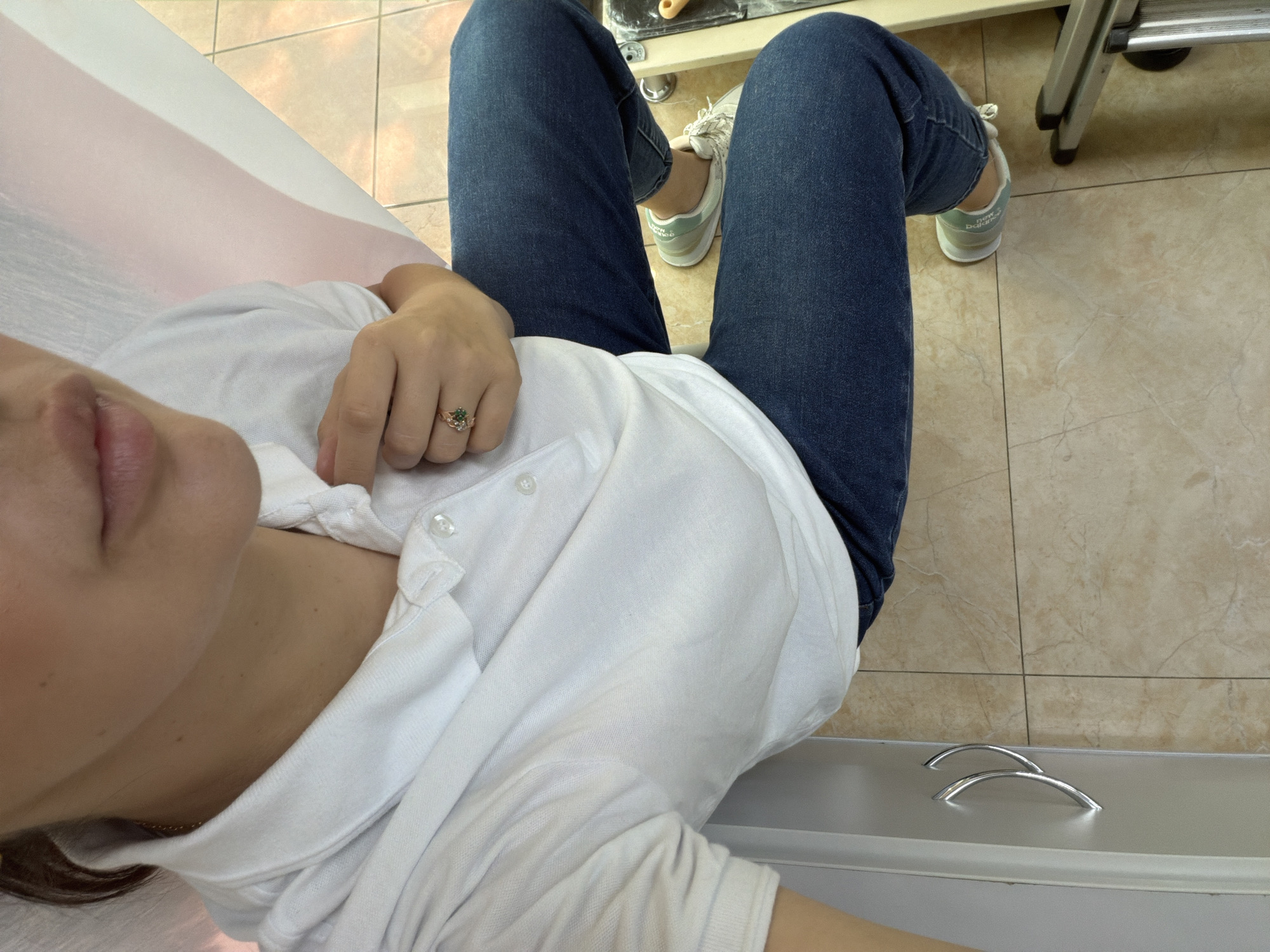 A close-up, overhead shot captures a person from the chest down, seated, in what appears to be a commercial establishment. The individual is wearing a white polo shirt and dark blue jeans. A ring with a green stone is visible on their right hand, which is resting on their chest. The person's feet are clad in light-colored sneakers with green accents. The background reveals tiled flooring and part of a light-colored cabinet with silver handles. The scene suggests an indoor setting, possibly a salon or spa, given the reflective surfaces and clinical appearance of the cabinet. The presence of a pink and white fabric strip, likely a curtain or drape, introduces a soft element to the otherwise sterile environment. There is no direct indication of the city of Sochi, Russia, in the image itself.