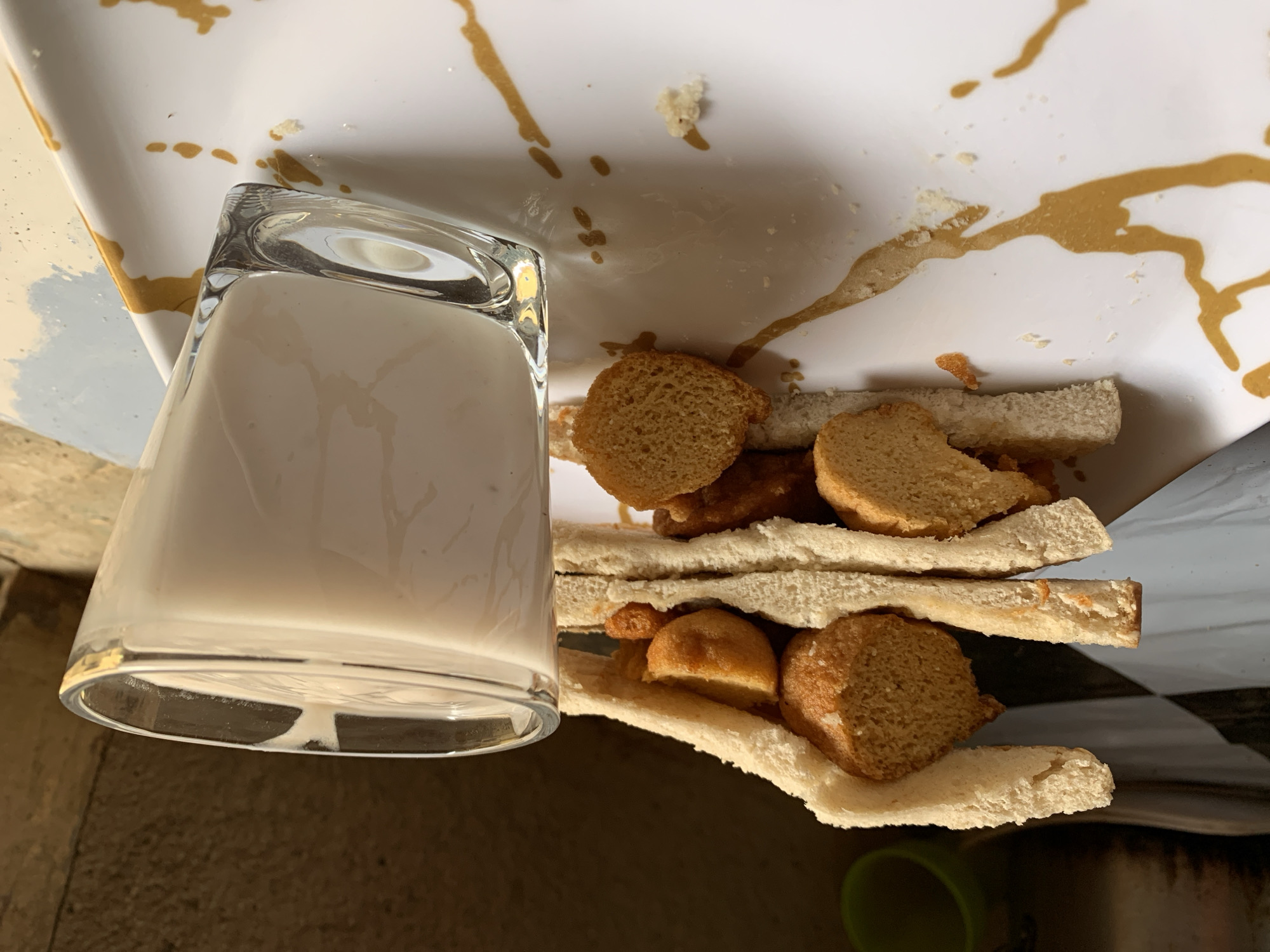 A glass of white liquid sits beside a plate containing pieces of bread with a filling, possibly a sandwich, on a surface in Rafin Par, Nigeria. There are brown stains on the plate.