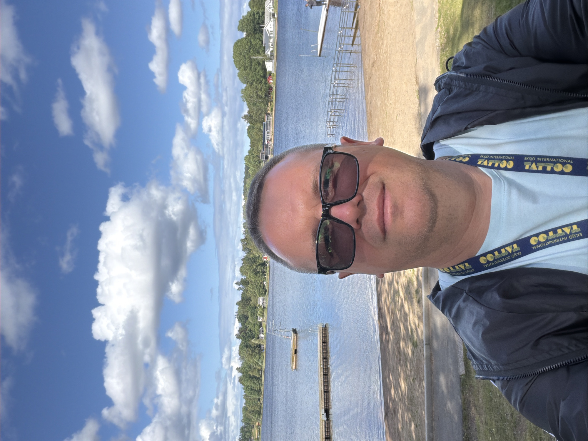 A selfie is taken by a man wearing glasses and a light blue t-shirt under a black jacket. He is standing on a sandy beach with a lake behind him. The lake has several wooden docks and a diving platform. In the background, there is a treeline with houses and buildings, and a blue sky with white clouds. The man is wearing a lanyard with the text "EKSIÖ INTERNATIONAL TATTOO EXPO" in yellow and blue. Based on the text on the lanyard, the photo was likely taken in Eksjö, Sweden.