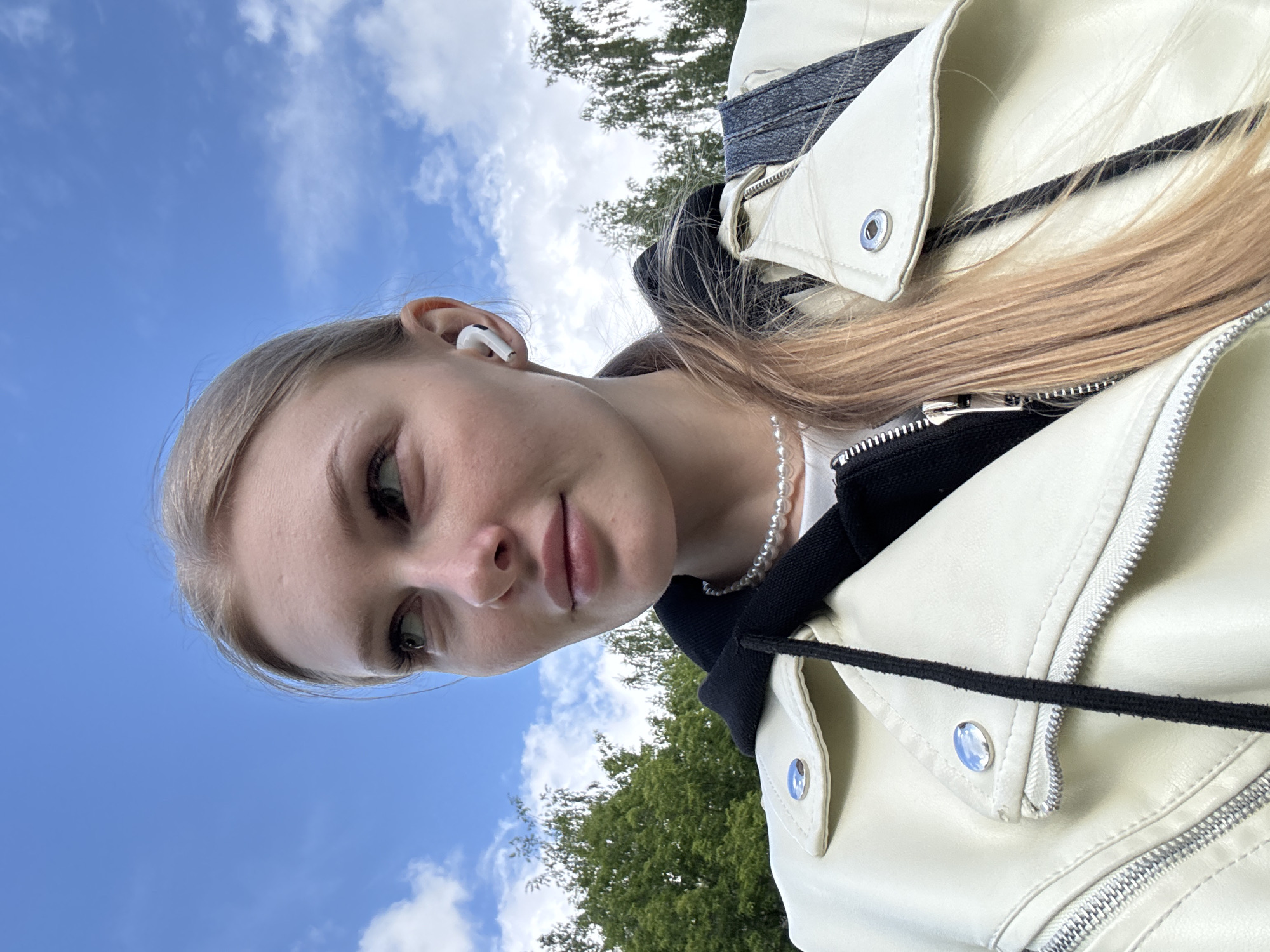 The image depicts a close-up, low-angle outdoor portrait of a young woman. She has fair skin, light-colored hair, and light-colored eyes, with visible eyelashes. Her lips are full, and she has a gentle, slightly smiling expression, looking towards her left (viewer's left). A white wireless earbud is visible in her right ear. She is wearing a cream-colored moto-style jacket with silver snaps/buttons and metallic zippers, layered over a black hooded top with visible drawstrings. Around her neck is a necklace composed of small, white pearl beads. A section of a dark-colored strap, possibly from a backpack or bag, is visible over her right shoulder. The background consists of a clear blue sky with scattered white clouds and the green foliage of deciduous trees, suggesting a park or natural outdoor setting. The specific location within the city of Perm, Russia, cannot be determined from the visual information provided in the image.