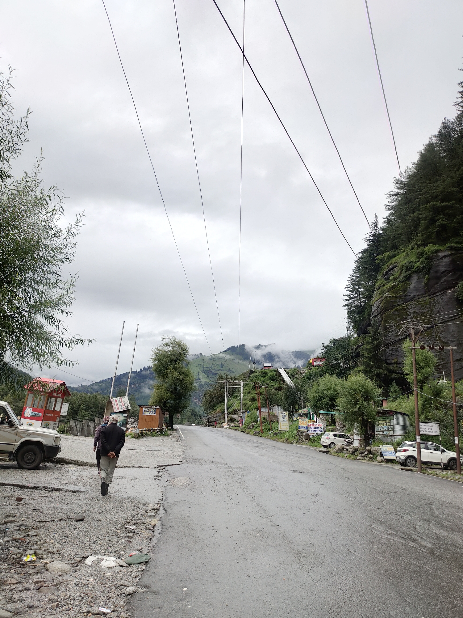 In Barheta, India, an overcast day presents a scene along a wet, asphalt road bordering a gravel shoulder. A man walks away from the viewer, his back to the camera, wearing a dark jacket and light-colored pants. To his left, a beige SUV is parked. Further down the road, a small red and white structure with a peaked roof stands on the left, and a weathered wooden booth is positioned on the right. A mountainous landscape with verdant slopes and misty peaks forms the backdrop. Power lines crisscross the sky. Trees line the right side of the road, with a steep, rocky cliff face partially covered in foliage. Several white cars are parked along the right side of the road.