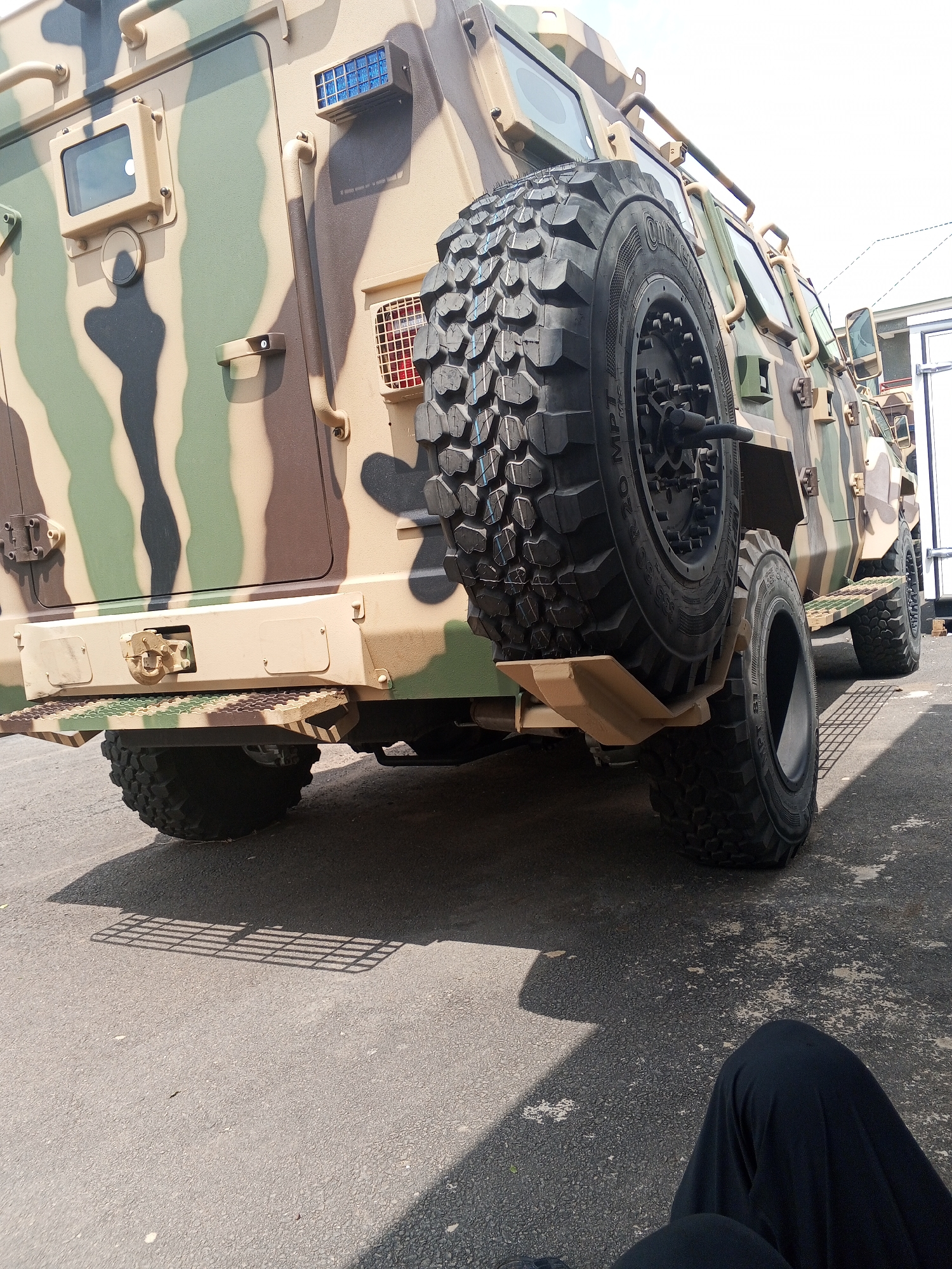 A close-up view of the rear of a camouflage military vehicle with a large spare tire mounted on the back. The vehicle is painted in a camouflage pattern of tan, green, and black. The large tire dominates the frame, displaying its tread pattern and size markings. Parts of a person's black clothing are in the lower right corner. The vehicle is situated on asphalt. The location is Aso, Nigeria.