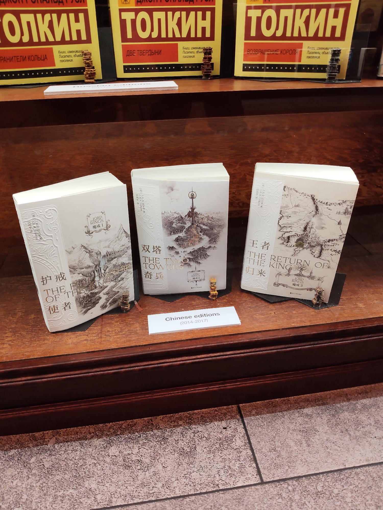 The image shows a display of three Chinese editions of J.R.R. Tolkien's "The Lord of the Rings" books. The books are displayed upright on black stands on a wooden surface. The titles are visible in both Chinese and English. The first book on the left has the title "The Fellowship of the Ring" (护戒使者). The middle book is "The Two Towers" (双塔奇兵). The book on the right is "The Return of the King" (王者归来). Each book has intricate embossed designs on the cover and a small, golden, miniature statue of a hobbit in front of it. In the background, there are three Russian editions of Tolkien's books, with yellow covers and Russian text. A white sign in the foreground reads "Chinese editions (2014-2017)". The overall setting appears to be a bookstore or library display. The location is Singapore, Singapore.