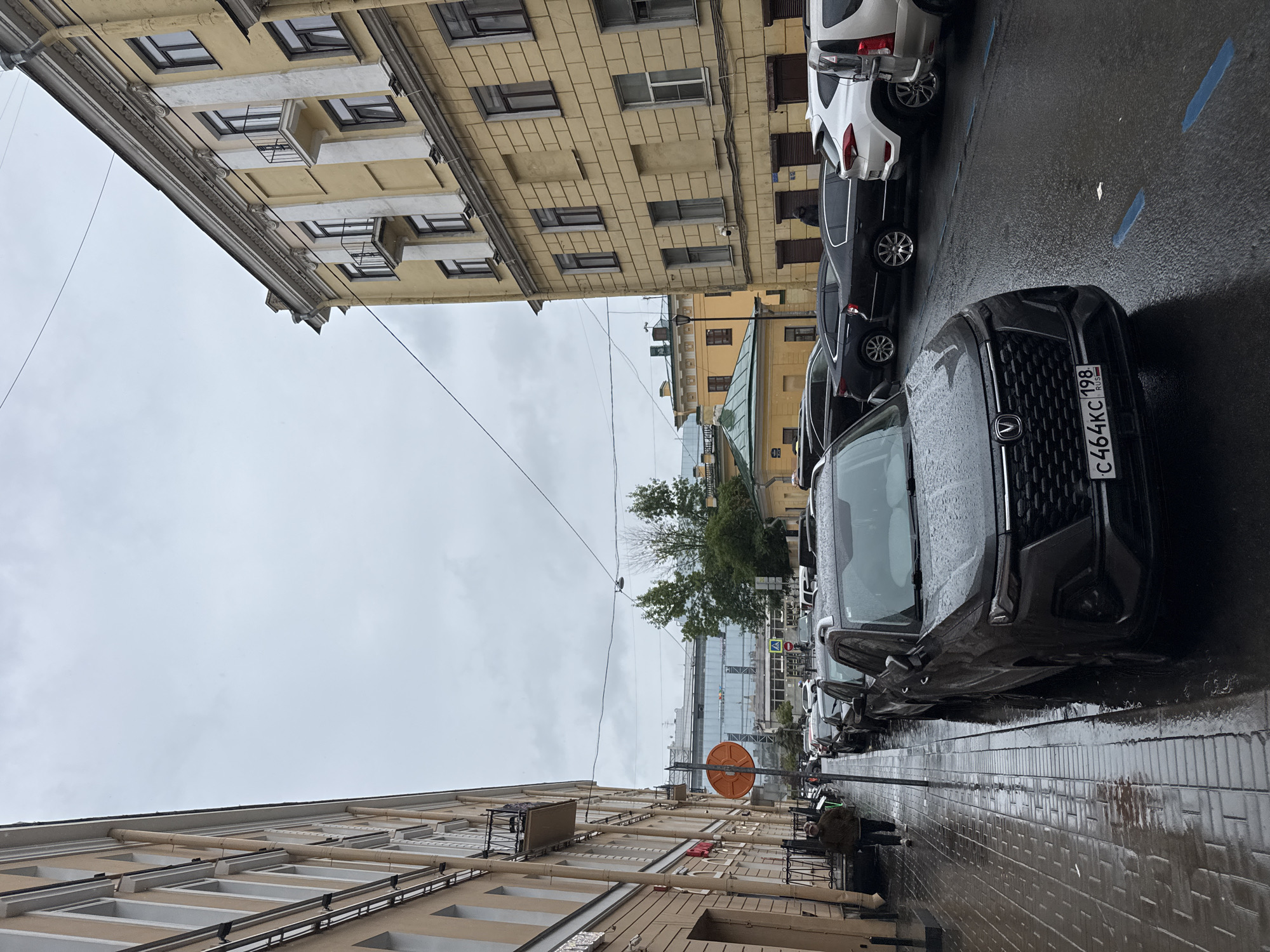 This is an eye-level, medium shot of a wet street in Saint Petersburg, Russia, on an overcast day. Cars are parked on the right side of the road, and pedestrians walk on the left sidewalk. The architecture suggests an urban setting, with tall, multi-story buildings lining both sides of the street. One building on the right is yellow with white decorative elements and balconies. The building on the left is a lighter color and also has balconies. The street is paved with cobblestones and has puddles from recent rain. A dark gray car with a Russian license plate is in the foreground. In the background, a large modern building with glass windows is visible. A person in a brown jacket is walking on the left sidewalk.