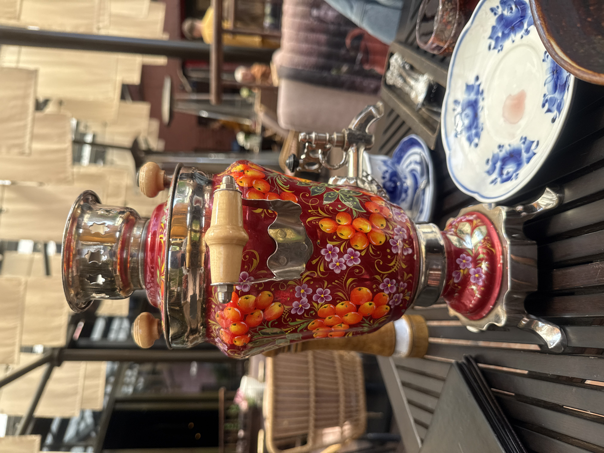 The image depicts a close-up of a decorative, red samovar with painted orange berries and purple flowers, positioned on a dark wooden table. The samovar has silver accents and wooden handles. To the left of the samovar, a stack of black menus is visible. To the right, a white saucer with blue floral patterns holds a small pinkish stain. In the background, obscured by the samovar and the foreground elements, a dimly lit outdoor seating area with wicker chairs and tables can be seen. The ambient lighting suggests it is daytime. A person in a yellow top is visible in the far background, out of focus. The scene appears to be an outdoor cafe or restaurant. Given the context of a samovar being prominently displayed, this scene is likely located in Russia. While the image doesn't provide specific visual cues to pinpoint Vladivostok, samovars are a common element in Russian hospitality and are frequently found in cafes and restaurants across the country.