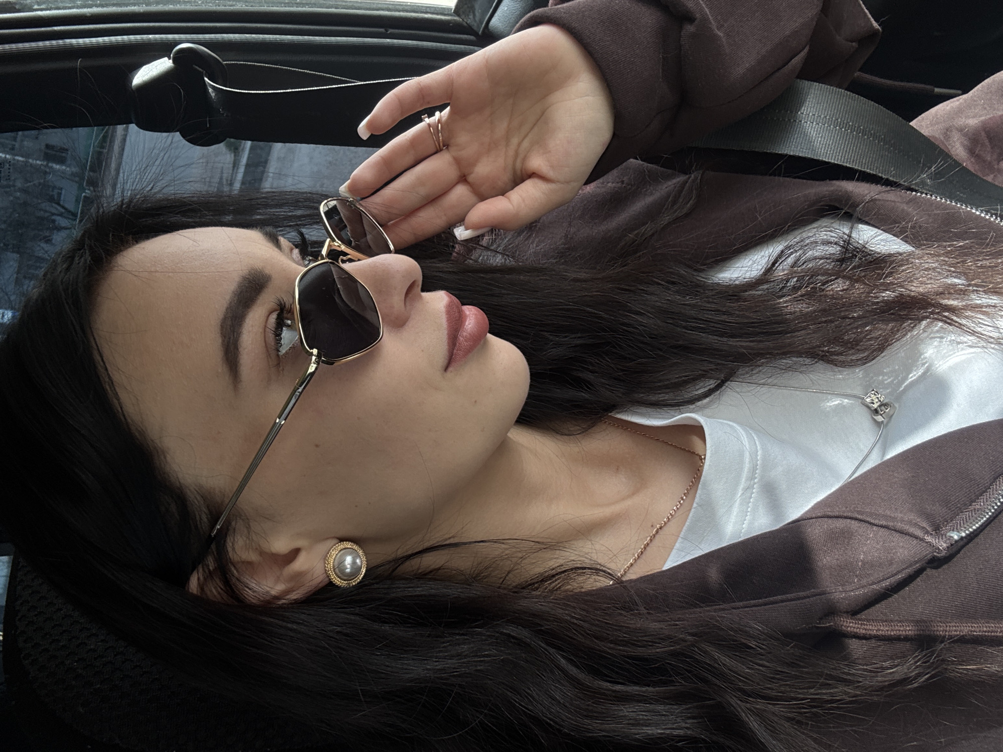 A person is lying down, potentially inside a vehicle, with their head resting on an unseen surface. They are wearing sunglasses with gold frames and a brown jacket over a white shirt. A gold necklace with a small pendant is visible around their neck. A ring is on one of their fingers. The scene suggests a relaxed or casual setting. No elements explicitly place the scene in Chaikovsky, Russia.