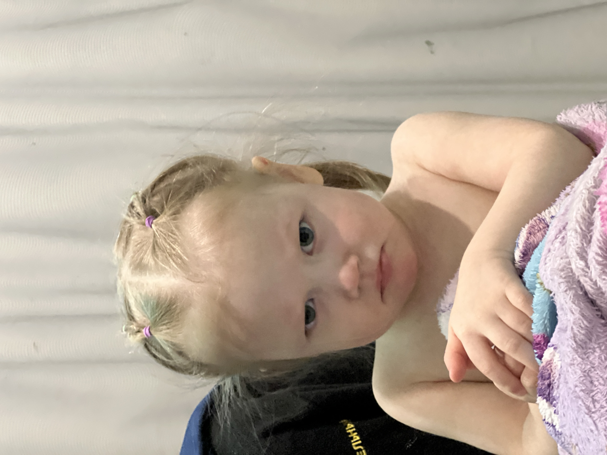 A young child with light blonde hair styled in pigtails secured with purple bands is partially wrapped in a purple and blue fuzzy blanket. The child is looking towards the camera with a slight frown. The background appears to be a plain wall. The lower portion of the image reveals a dark blue or black fabric. The photograph was taken indoors, but there is no other information to place it in Perm, Russia.