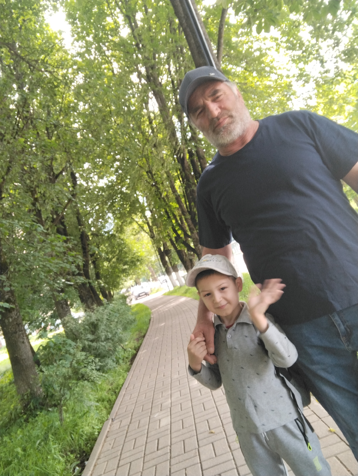 In Kingisepp, Russia, a man and a boy stand on a paved walkway lined with trees. The man, wearing a dark t-shirt and a baseball cap, has grey hair and a beard and is looking towards the camera. The boy, dressed in a grey shirt and pants with a baseball cap, is standing in front of the man, holding his hand and waving. The walkway is made of light-colored paving stones and curves to the right. Green grass and trees are visible on either side of the walkway, and in the background, there are more trees and what appears to be a street with vehicles. The lighting suggests it is daytime, possibly overcast or late afternoon, with sunlight filtering through the trees.