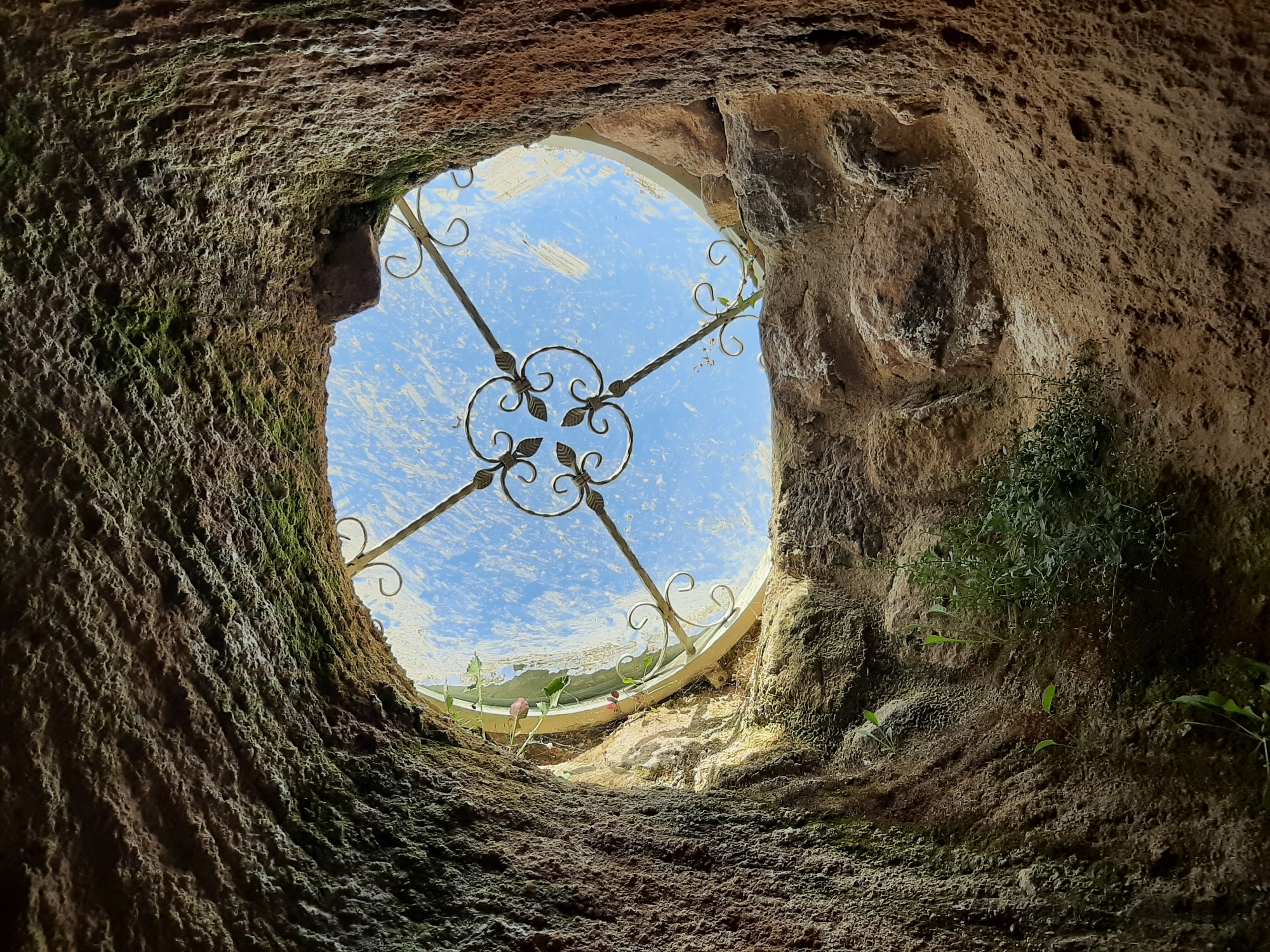A low-angle, wide shot captures a circular opening in a textured, earthy structure, looking up towards a bright blue sky. The opening is framed by rough, uneven walls of reddish-brown earth, with patches of green moss clinging to the left side. A decorative, wrought-iron grate with floral elements is visible across the opening, set against a hazy, frosted glass or plastic cover that allows light to filter through. The grate is attached to a white, circular frame that encloses the opening. Small green plants with delicate pink buds are growing from the edges of the opening, near the grate and frame. The overall impression is of a rustic, natural setting with a touch of human craftsmanship.