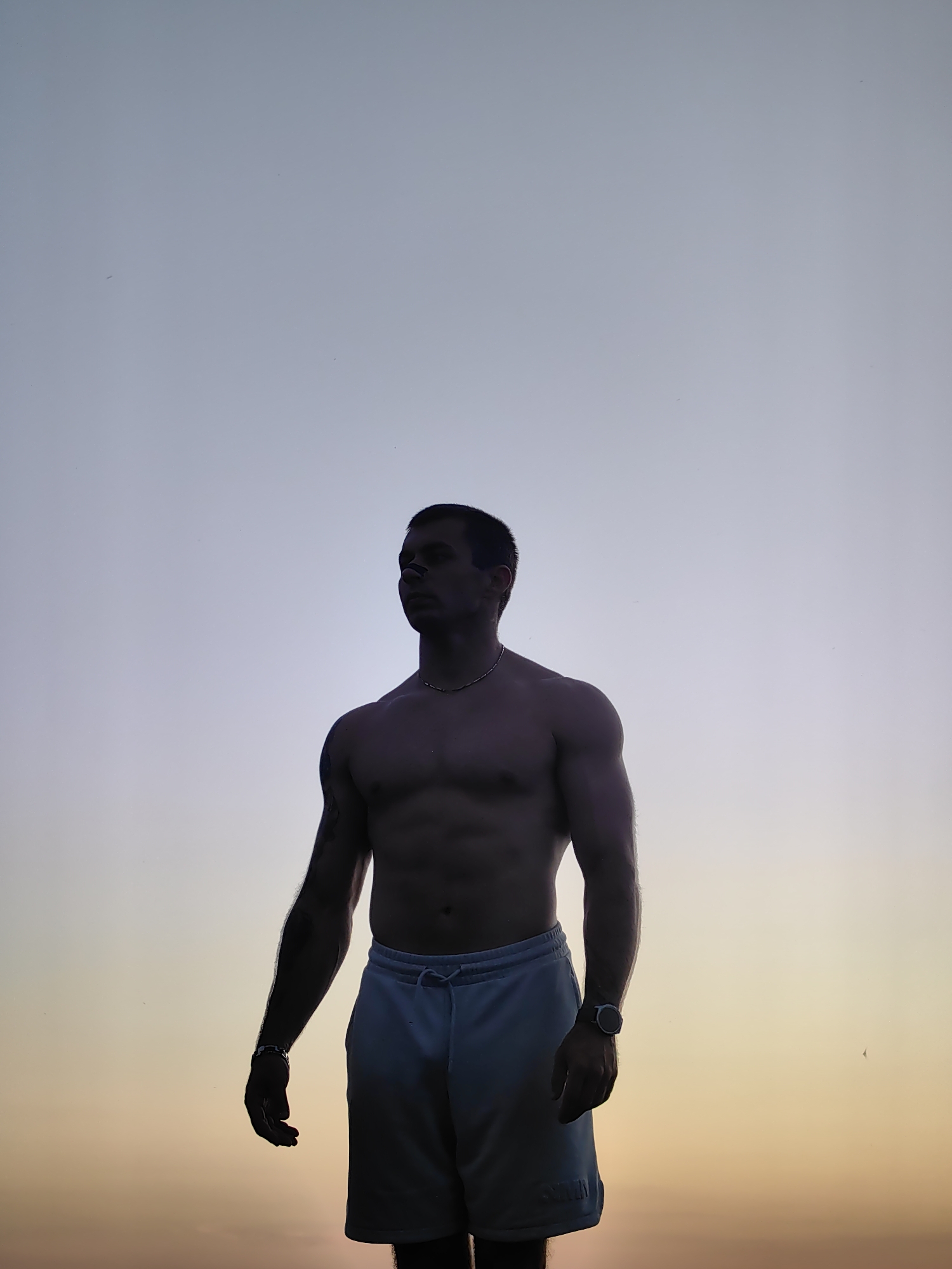 A shirtless man with dark hair stands in silhouette against a gradient sky that transitions from light blue at the top to a warm yellow and orange hue at the bottom, suggesting it is either sunrise or sunset. He is wearing light-colored shorts and a necklace. A watch is visible on his left wrist. The man's body is toned, and he is looking to his left. There are a few small, dark specks in the sky that could be birds or insects. The location is identified as Varna, Bulgaria, but no specific landmarks are visible to confirm this.