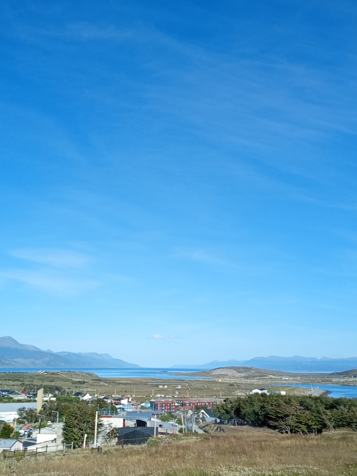 A panoramic view showcases a clear blue sky dominating the upper portion of the image. Below, a landscape unfolds with rolling hills covered in dry grasses leading down to a town with colorful buildings nestled among trees. Further in the distance, a body of water stretches towards the horizon, framed by distant mountains. The scene captures a sense of vastness and peacefulness.