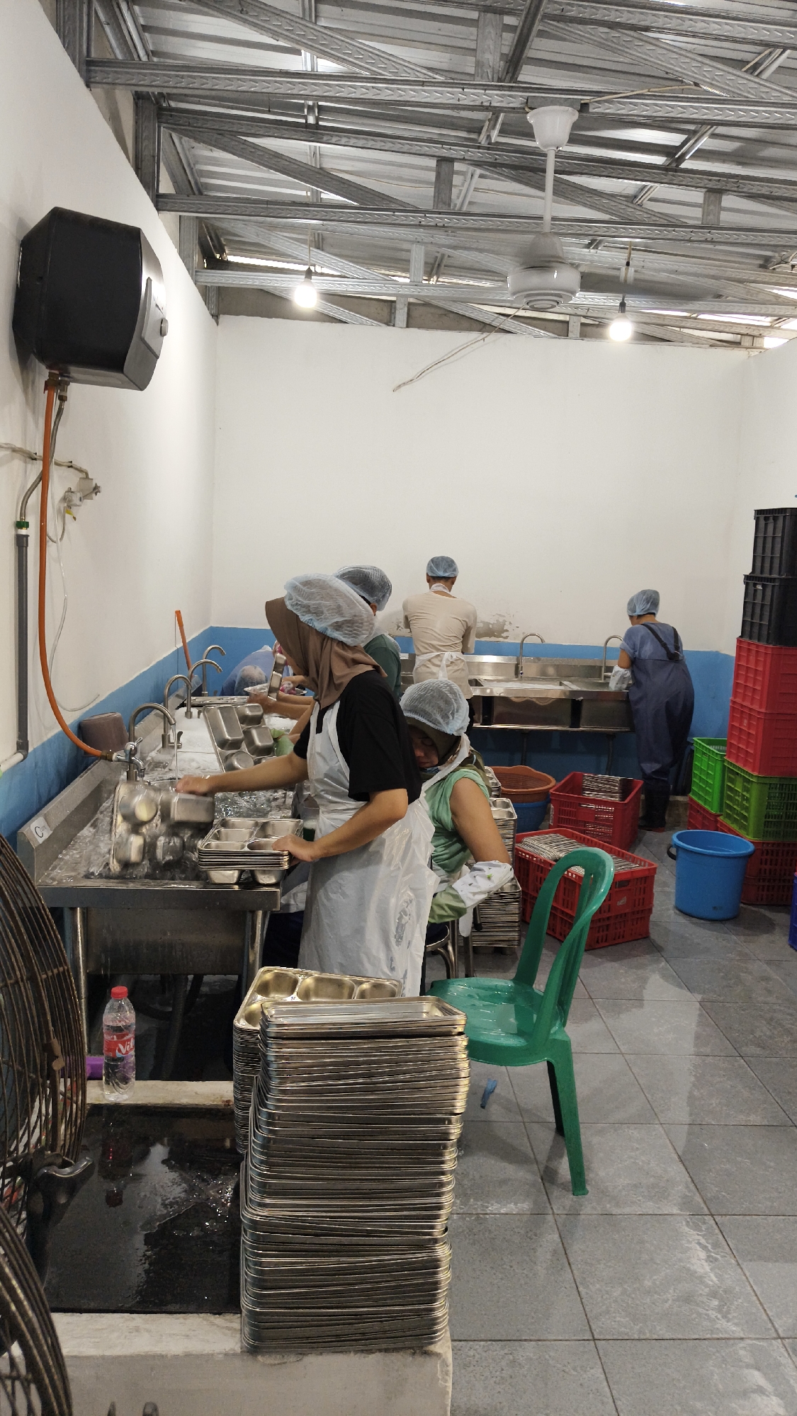 The image depicts an interior scene, likely a food preparation or washing area, within Sambipondok, Indonesia. The environment appears to be a commercial or industrial kitchen with white tiled walls and a grey tiled floor. A metal ceiling structure with visible beams and light fixtures is overhead, along with a white ceiling fan.
In the foreground, a stack of metal food trays is prominently displayed on a ledge. To the left, a large black fan with a circular grill is partially visible. A red and white plastic water bottle is placed on a wet surface next to the fan.
The main activity involves several individuals engaged in washing dishes. In the center, a woman wearing a hijab and an apron is actively washing trays in a large stainless steel sink with multiple faucets. She is holding a stack of trays. To her right, another individual, also wearing a hairnet, is seated and appears to be assisting or organizing washed items. Further in the background, two other individuals are visible near sinks, also seemingly involved in the washing process. They are all wearing hairnets and aprons or protective clothing, indicating a focus on hygiene.
The background features several stacks of colorful plastic crates (red, green, and black) and a blue bucket, suggesting organized storage or transport of items. The overall impression is one of a functional, albeit somewhat basic, food service operation.