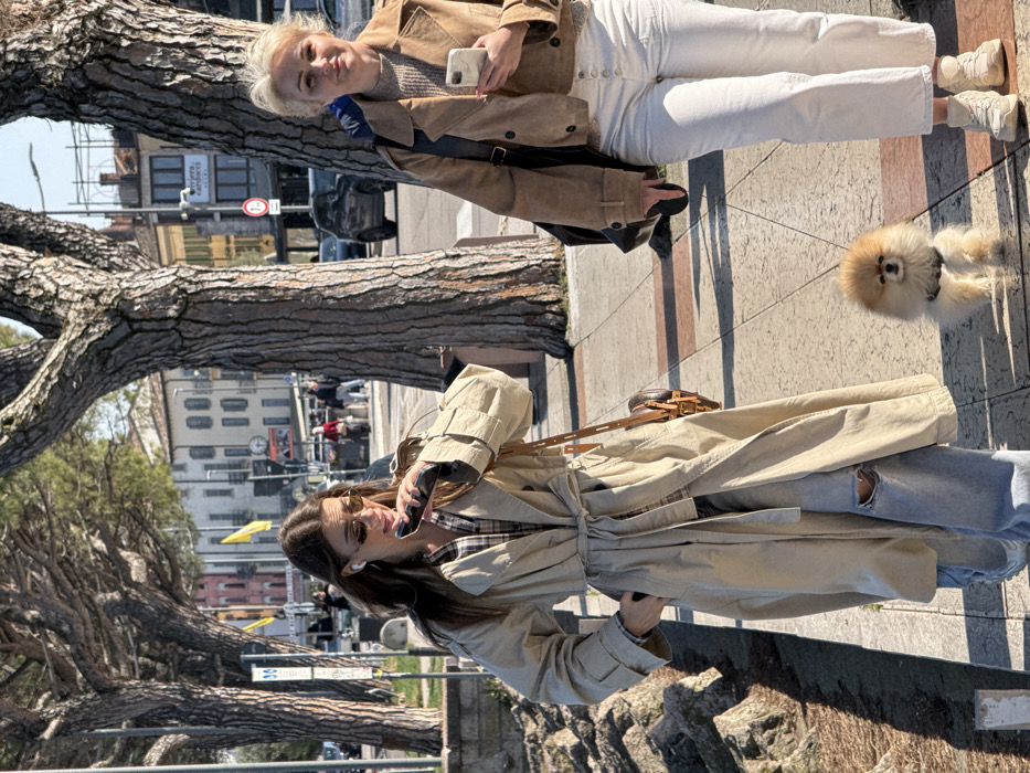 The image captures two women and a dog walking on a sunlit, tree-lined sidewalk in Peschiera del Garda, Italy. The paved sidewalk features light-colored square tiles, bordered by a low stone wall on the left and a series of mature trees with coarse bark. In the background, a street is visible with vehicles and multi-story buildings, some with light-colored facades and balconies. A street sign in the background clearly indicates "Via Giuseppe Carducci."
The woman on the left, with long brown hair, wears small oval sunglasses, white wireless earbuds, a light beige trench coat tied at the waist, a plaid shirt, and ripped light blue jeans. She is holding a dark smartphone to her mouth, appearing to be speaking into it or recording a voice message, and has her right hand in her coat pocket. A small brown crossbody bag with gold hardware is slung over her left shoulder.
The woman on the right, with blonde hair, wears a light brown cropped jacket, a light brown knit sweater, high-waisted white wide-leg jeans with visible buttons, and white sneakers. She holds a white smartphone in her left hand and a dark shoulder bag on her right side. She is looking towards the camera with a neutral expression.
A small, fluffy, light brown Pomeranian-type dog, wearing a dark harness, walks on the sidewalk between the two women, slightly ahead of the woman on the right. The scene is illuminated by bright, direct sunlight, casting clear shadows on the ground.
