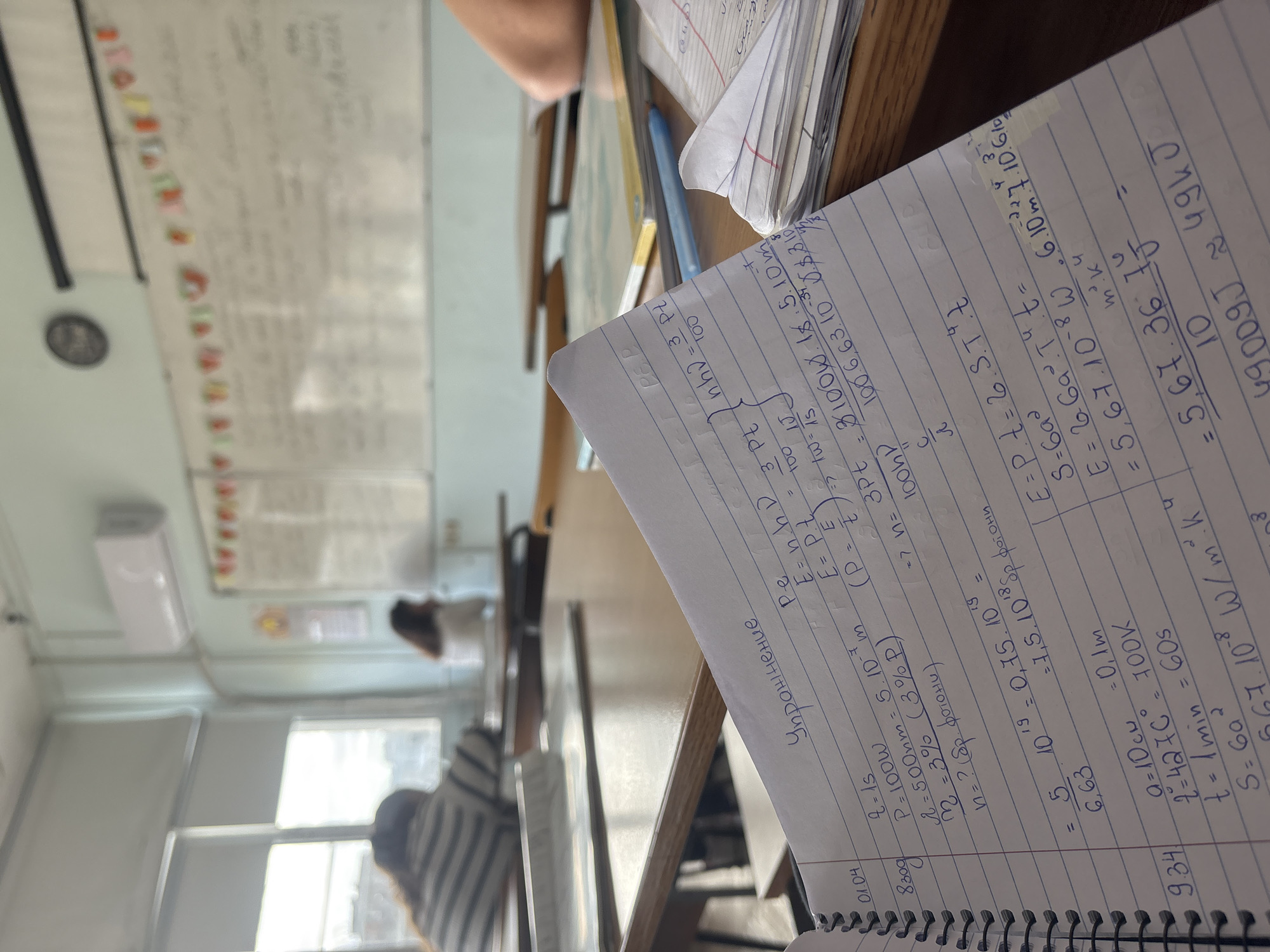 A close-up shot focuses on a notebook page filled with handwritten physics calculations, seemingly related to energy and radiation. The calculations involve variables like power (P), wavelength (λ), temperature (T), and Stefan-Boltzmann constant (σ), suggesting topics such as blackbody radiation or heat transfer.
The notebook sits on a wooden desk, partially obscured by other papers and a pen. In the background, a classroom setting is visible, complete with a whiteboard covered in writing, a radiator, and students at desks. The environment suggests an educational context, possibly a physics class in session.
While the location appears to be a classroom setting, there is no visual information that suggests the image was taken in the city of Trilistnik, Bulgaria.