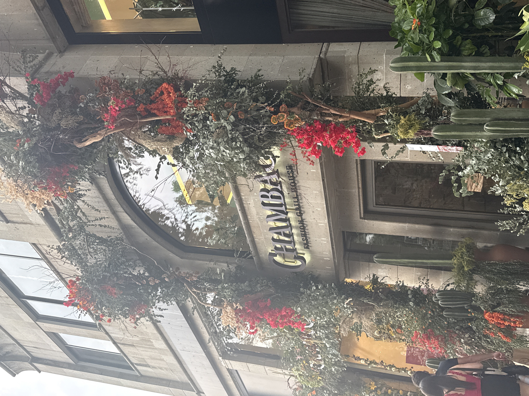 The image shows the exterior of "CHAMBA", a fashion grill house in Mexico City, Mexico. The building is made of grey stone. The entrance is decorated with green and red floral arrangements and cacti. There is a partial view of people in the lower right corner.