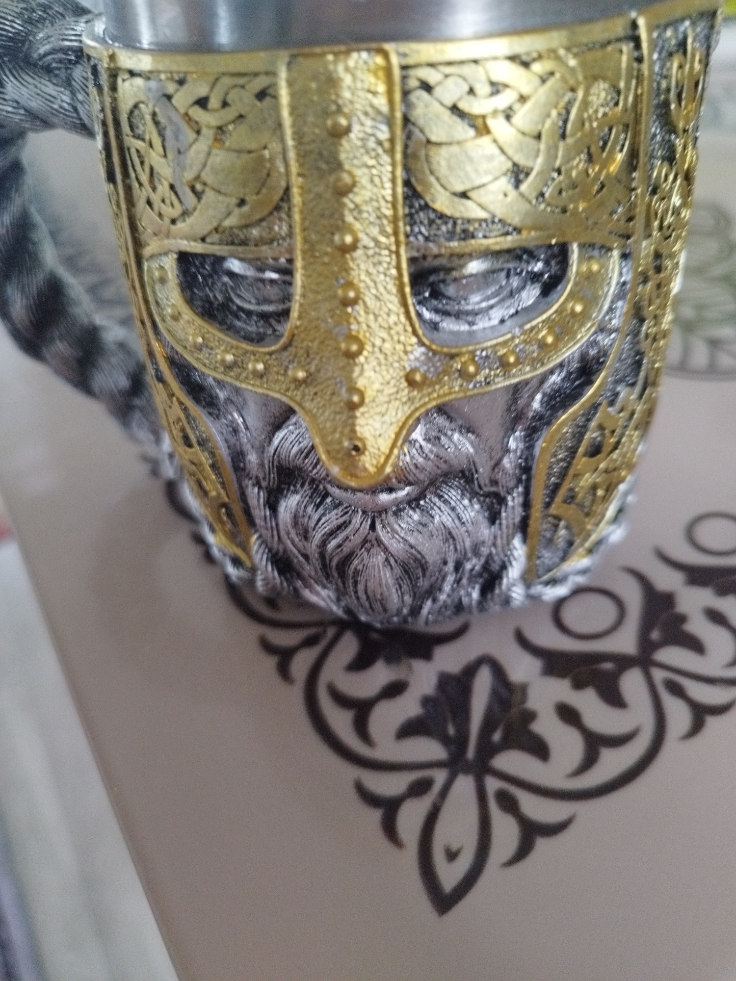 A close-up, eye-level, indoor shot shows a mug with a detailed, ornate design. The mug appears to be made of metal, with a silver-colored base and a gold-colored embellishment that resembles a Viking helmet with a beard. The helmet has intricate knotwork patterns in gold on the sides and around the eyeholes. The beard is textured to look like flowing silver hair. The mug is resting on a light-colored surface with a dark, decorative pattern. The background is out of focus. There are no people visible in the image.
