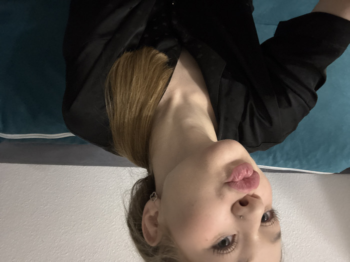 A young woman with fair skin, long blonde hair, and light eyes is centered. She is wearing a black top. Her lips are pursed slightly. A small nose piercing is visible on her left nostril. She wears a clover-shaped earring. She's situated on a blue and white surface, possibly a bed or couch. The image is oriented at an angle.