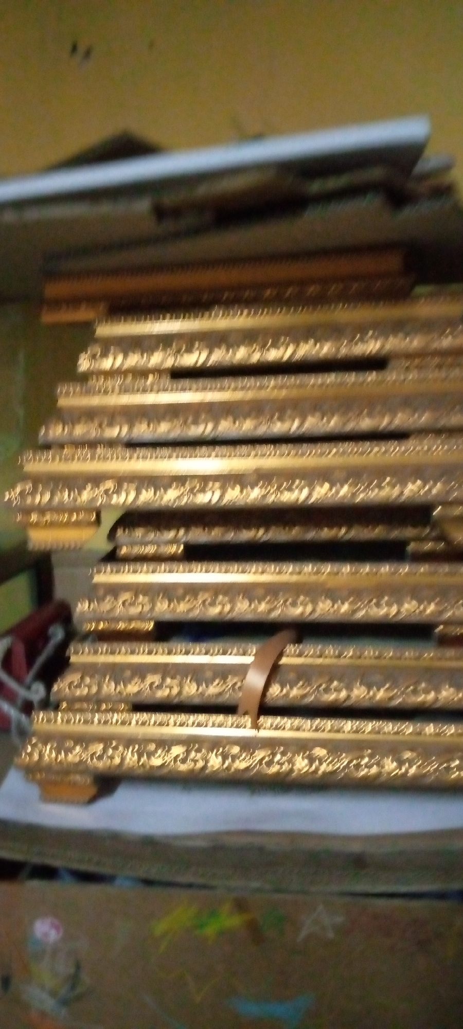 The image shows a stack of golden ornate picture frames. The frames are of various sizes and appear to be stored horizontally, one on top of another. Some of the frames have intricate carvings and decorative patterns. The background is out of focus, but suggests an indoor setting with a yellow wall. A brown strap is visible across some of the frames. The lower part of the image shows the top of a cardboard box with colorful drawings, possibly made with chalk or crayons. This scene is located in Sumberwono, Indonesia.