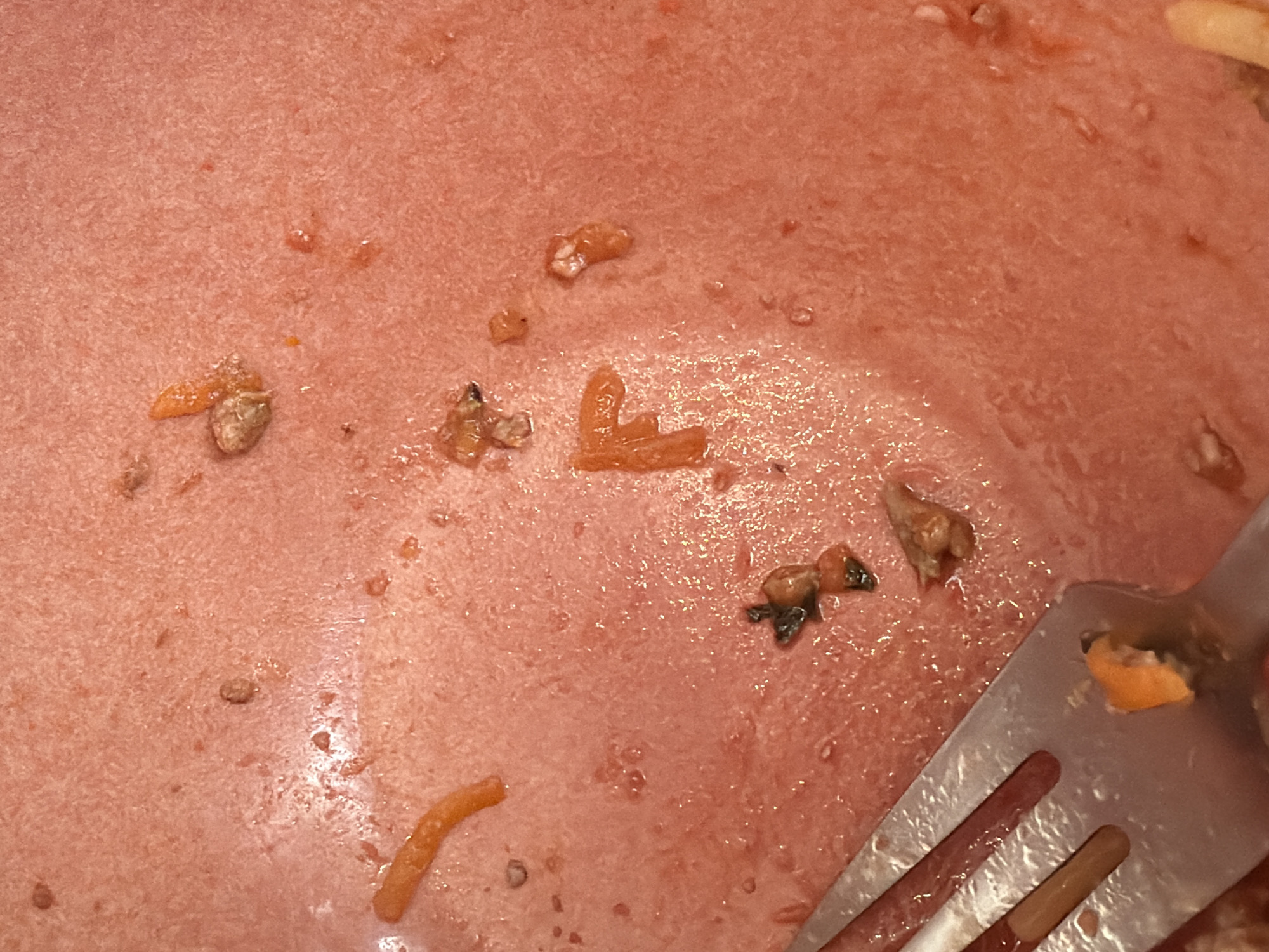 The image shows a close-up view of a plate after a meal, with remnants of food and a fork. The plate appears to contain leftover pasta with a red sauce, possibly tomato-based. Small pieces of pasta, meat, and vegetables are scattered across the surface. A metal fork is visible on the right side of the frame, with some food residue clinging to its tines. The overall impression is of a partially eaten meal on a plate. There is no evidence to indicate the location is Budva, Montenegro.