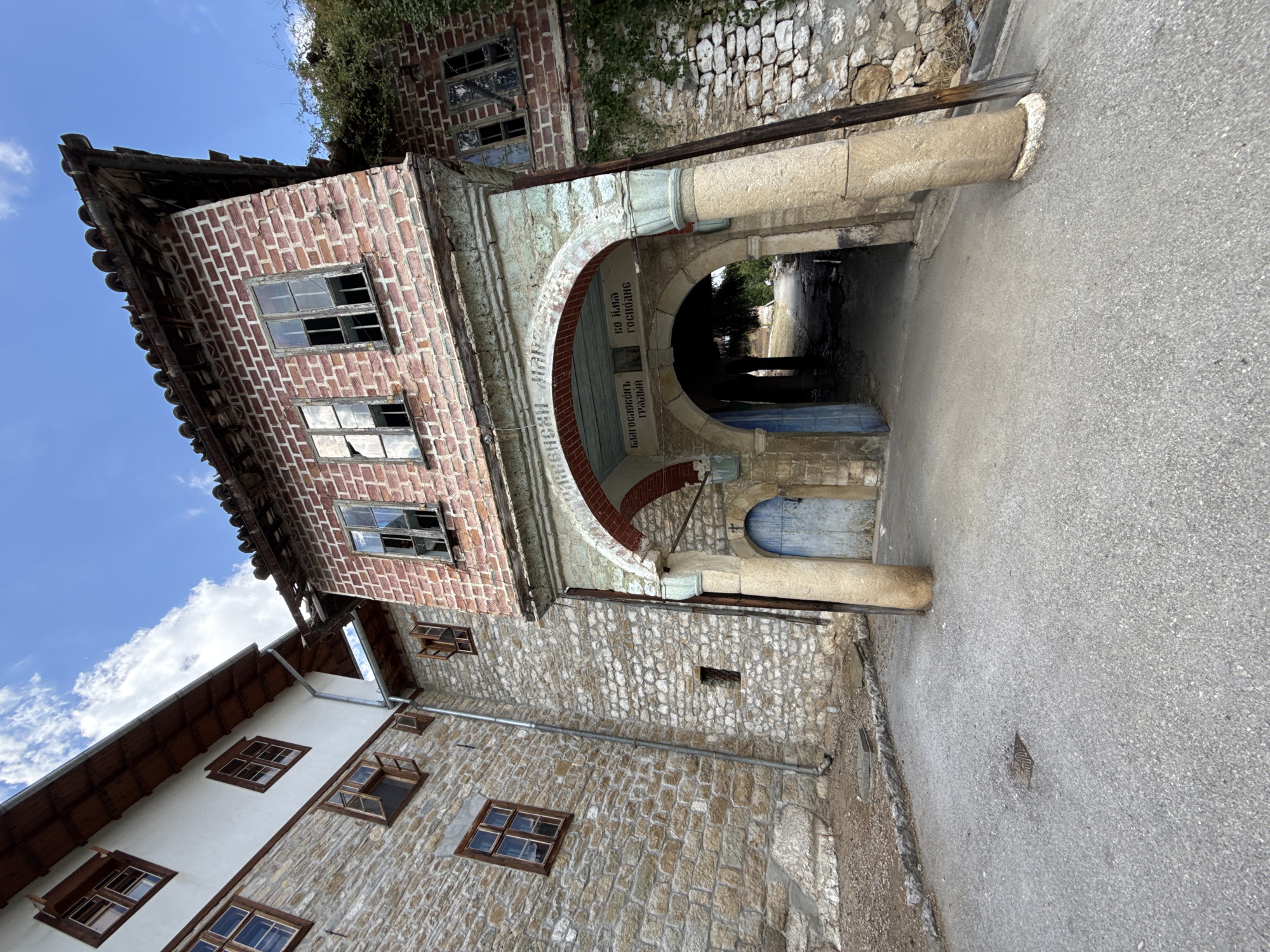 The image shows an arched gateway made of stone and brick, leading into a dark passage. Above the archway, there is an inscription in Cyrillic script. The gateway is flanked by stone pillars and leads to a stone building with a tiled roof. To the left of the gateway, a larger stone building with white plastered walls and wooden windows is visible. The ground in front of the gateway is paved with asphalt. The scene appears to be in Samovodene, Bulgaria, likely a historic or religious site. The architectural style suggests a traditional Bulgarian building. The sky is cloudy, and the light suggests it is daytime. There are no people or animals visible in the image.