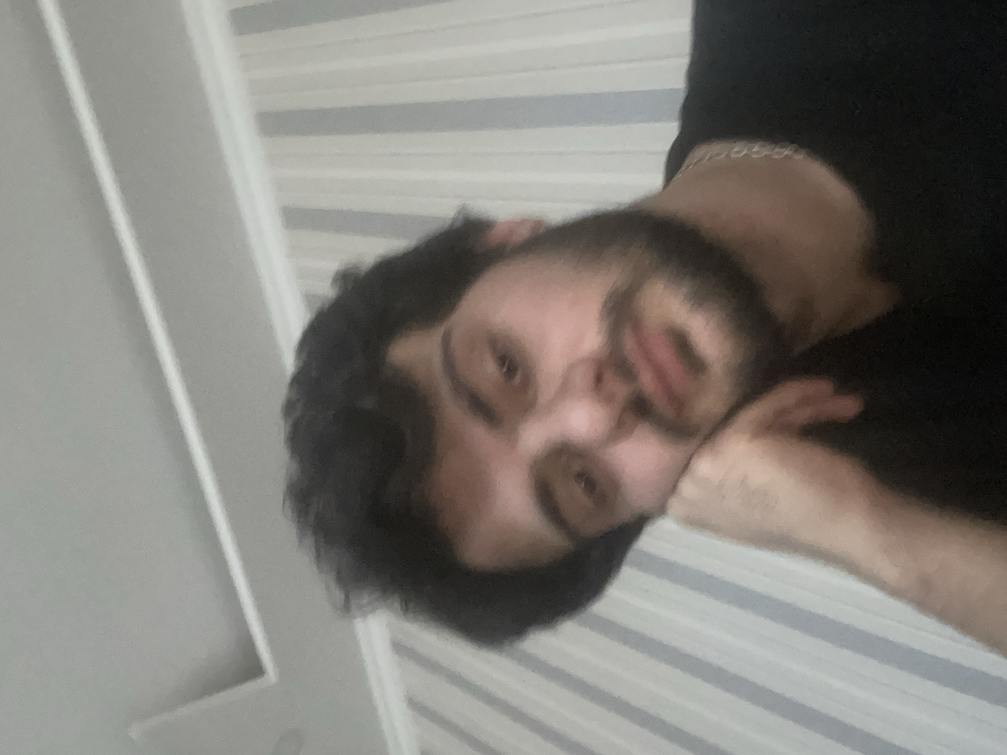 The image is a close-up, over-the-shoulder selfie of a young man with dark, curly hair and a beard, resting his cheek on his hand. He is wearing a black t-shirt and a silver chain necklace. The background consists of a wall with vertical light blue and white striped wallpaper and a white ceiling with recessed lighting. The man's expression appears thoughtful or tired. The scene is indoors, likely a residential or hotel room, and there are no visible indicators to place it specifically within Tbilisi, Georgia.