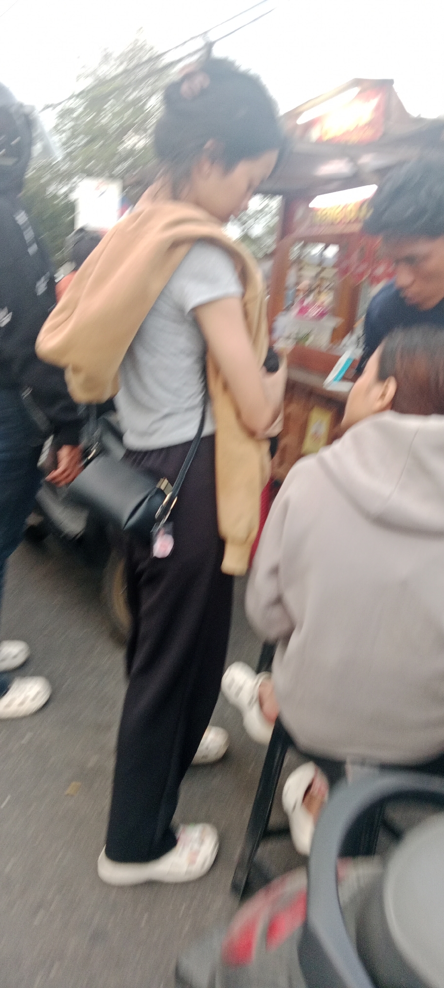 A slightly blurred, medium shot captures a group of people standing near a food cart in Ngemplak, Indonesia. The scene is outdoors, possibly a street market. In the foreground, a woman wearing black pants and white patterned clogs is seen from the waist down. She has a tan sweater draped over her shoulders, a gray t-shirt, and a black purse with a gold buckle. Her hair is styled in a bun. To her right, a person in a light gray hooded sweatshirt is seated on a black stool, also wearing white clogs. Behind them, a food cart with red accents and bright signage is visible. To the left, a person wearing a black hooded jacket and blue jeans stands partially obscured, with their hand reaching out. The ground is paved, suggesting a street or walkway. The overall impression is of a casual, daytime gathering at a street vendor.