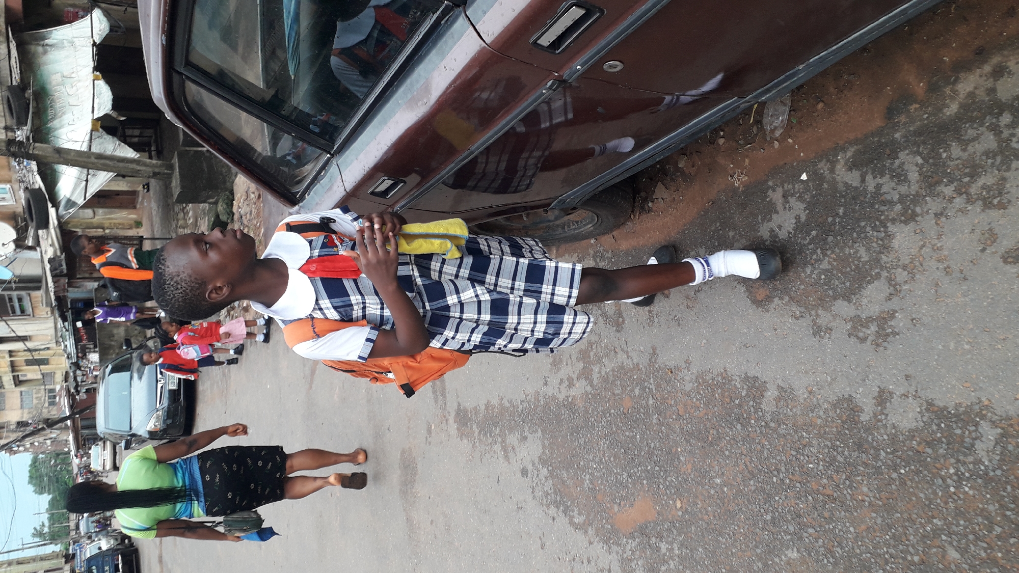 A young girl in a school uniform stands on a city street, next to a brown car, holding a yellow cloth. She is wearing a blue and white plaid dress, white socks, and black shoes, and she is wearing an orange backpack. A woman with long, dark hair and wearing a green top and black skirt walks on the street nearby. Cars are parked along the street, and other people are walking in the background. The photo was taken in the city of Onitsha, Nigeria. The street appears to be made of asphalt. The sky is visible in the top left corner.