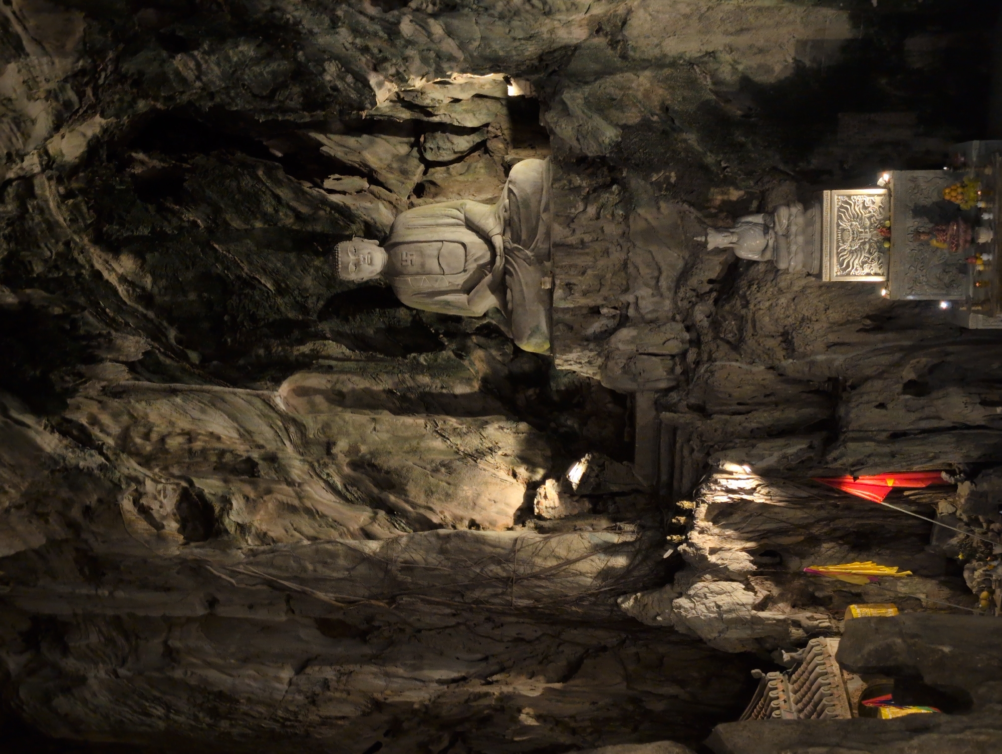 This image depicts the interior of a cave shrine in Đà Nẵng, Vietnam. A large, stone Buddha statue is prominently seated on a rocky platform in the center of the cave, bathed in artificial light. To the right of the main Buddha, a smaller, seated Buddha statue rests on an altar adorned with intricate carvings and lit by candles. In the foreground, on the left side of the image, a traditional Vietnamese temple roof structure is visible. Red and yellow flags are displayed, one of which is a red flag with a yellow star, likely representing Vietnam. The cave walls are rough and textured, with natural rock formations and some areas lit by spotlights, highlighting the spiritual and natural elements of the scene.