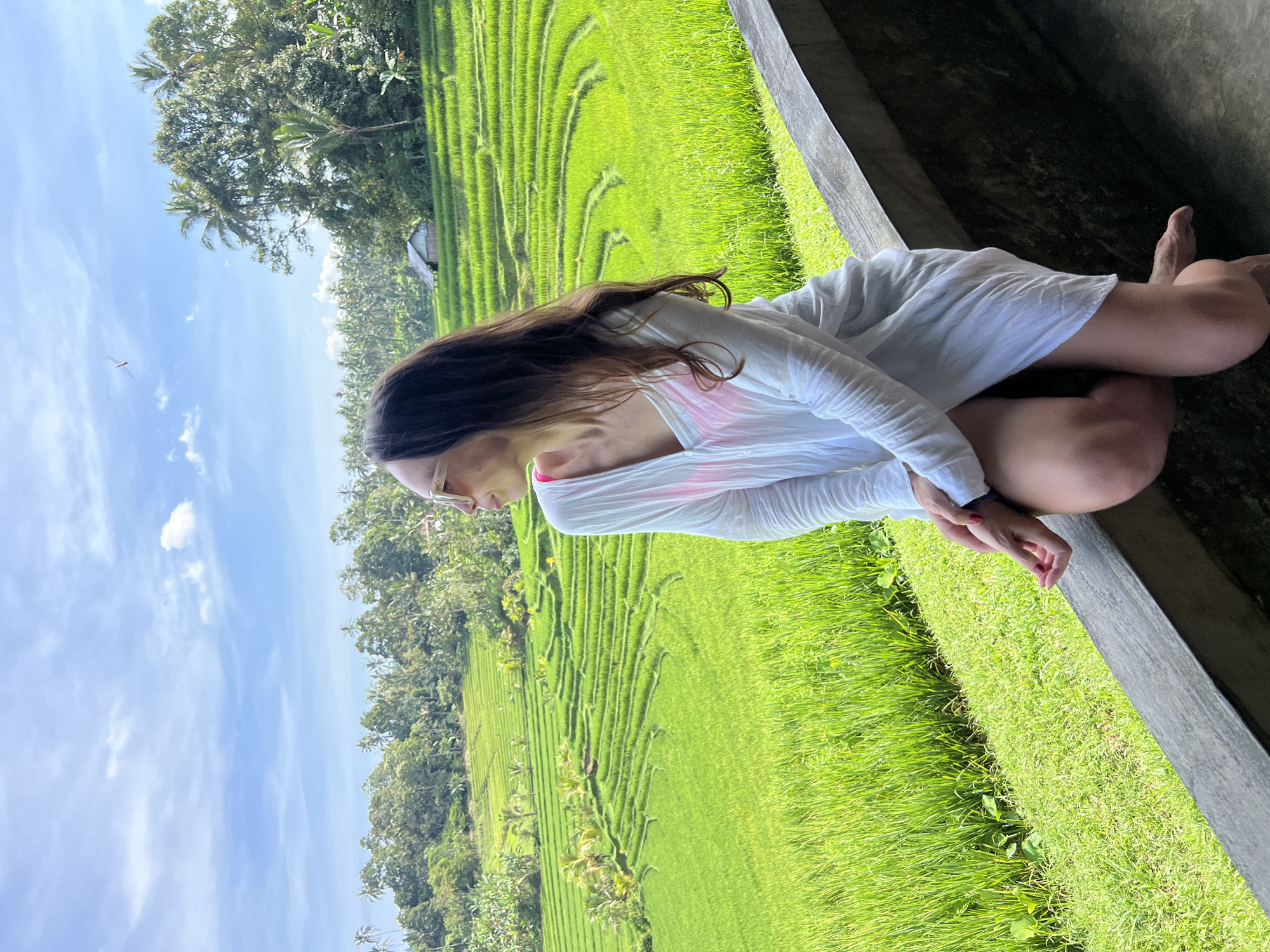 A woman is sitting with her legs crossed on a ledge overlooking a lush, green rice paddy field. She wears a white, long-sleeved shirt over a pink top. Her dark hair cascades down her shoulders, and she wears glasses. The background features terraces of rice paddies in Pupuan, Indonesia, with a clear sky dotted with a few clouds. Trees surround the rice paddies.