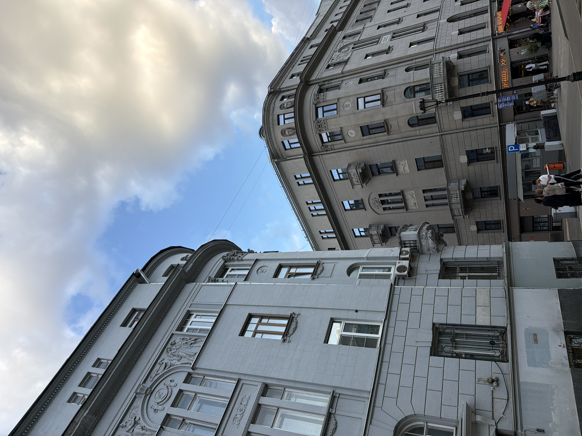 The image shows a low-angle view of two gray, ornate buildings against a sky with clouds and blue patches. The buildings have multiple windows, and some have decorative elements and balconies. On the right side of the image, there are signs, possibly for a restaurant called "ПИЦЦА 22 C". A street lamp is visible. People walk on the sidewalk. The location appears to be an urban setting.