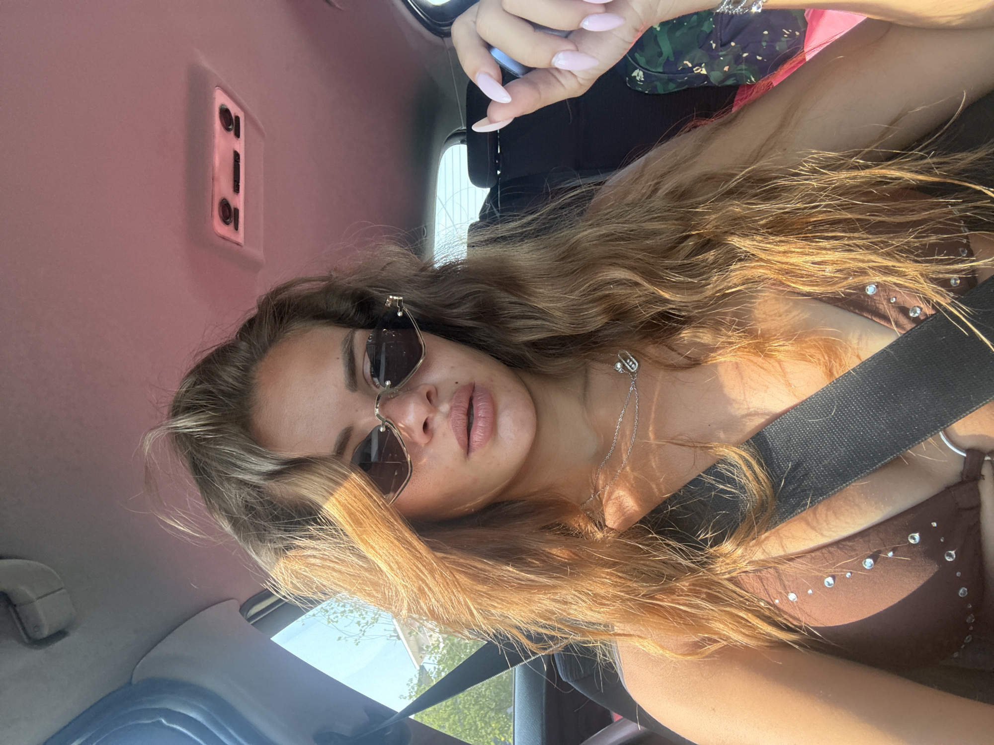 A woman with long, wavy, blonde hair is pictured in the front seat of a car, looking directly at the camera. She is wearing heart-shaped sunglasses and a brown bikini top adorned with rhinestones. Her left hand, with long, light pink painted nails, is visible near her face, holding a dark object. A black seatbelt is across her chest. The interior of the car has a pink ceiling lining and a light fixture on the roof. Sunlight streams in from the left, illuminating her hair and face. The background shows trees and a building, suggesting an outdoor setting, possibly in Katerini Municipality, Greece, during the daytime.
