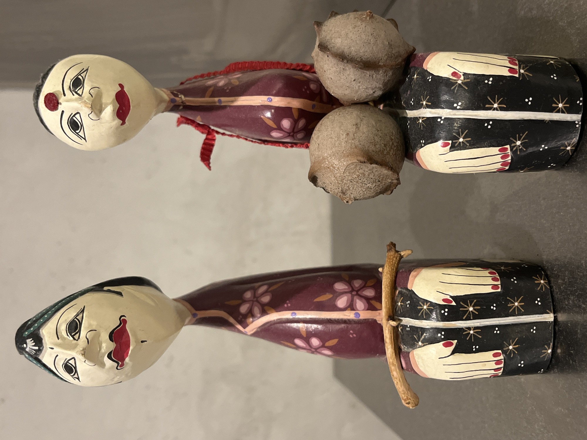 The image shows two hand-painted wooden dolls lying on a gray surface. The dolls have pale faces with painted features, including red lips, and are dressed in stylized clothing with floral patterns. The dolls are not located in Budva, Montenegro. They have no discernible relationship to the location.