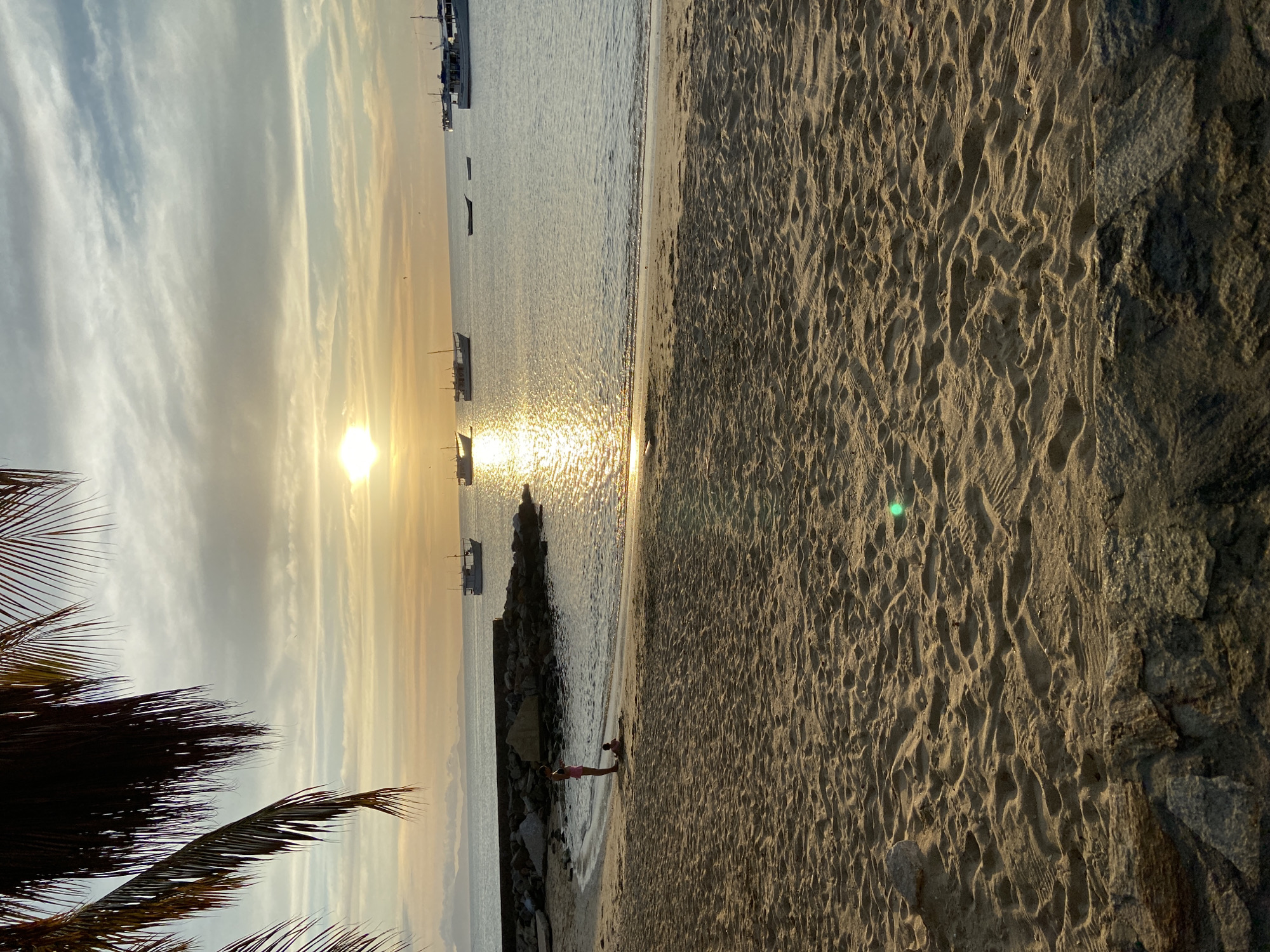 Here's a description of the image:
**Location:** The image appears to be taken on a beach in Juan Griego, Venezuela at sunset. This determination is based on the coastal setting, presence of boats, and the specific lighting conditions associated with sunset in a tropical location.
**Environment:** The scene is bathed in golden light from the setting sun. The sky is partly cloudy. The beach is sandy, with the texture indicating it may be coarse-grained. There are large rocks or a breakwater structure along the shore. Palm tree fronds are visible in the bottom left of the frame, indicating the presence of tropical vegetation.
**Subjects & Objects:** Several small boats are anchored in the water, silhouetted against the bright sunlight. Two people, possibly children, are standing in the shallow water near the rocks. The primary objects are natural elements: sand, water, rocks, and a palm tree.