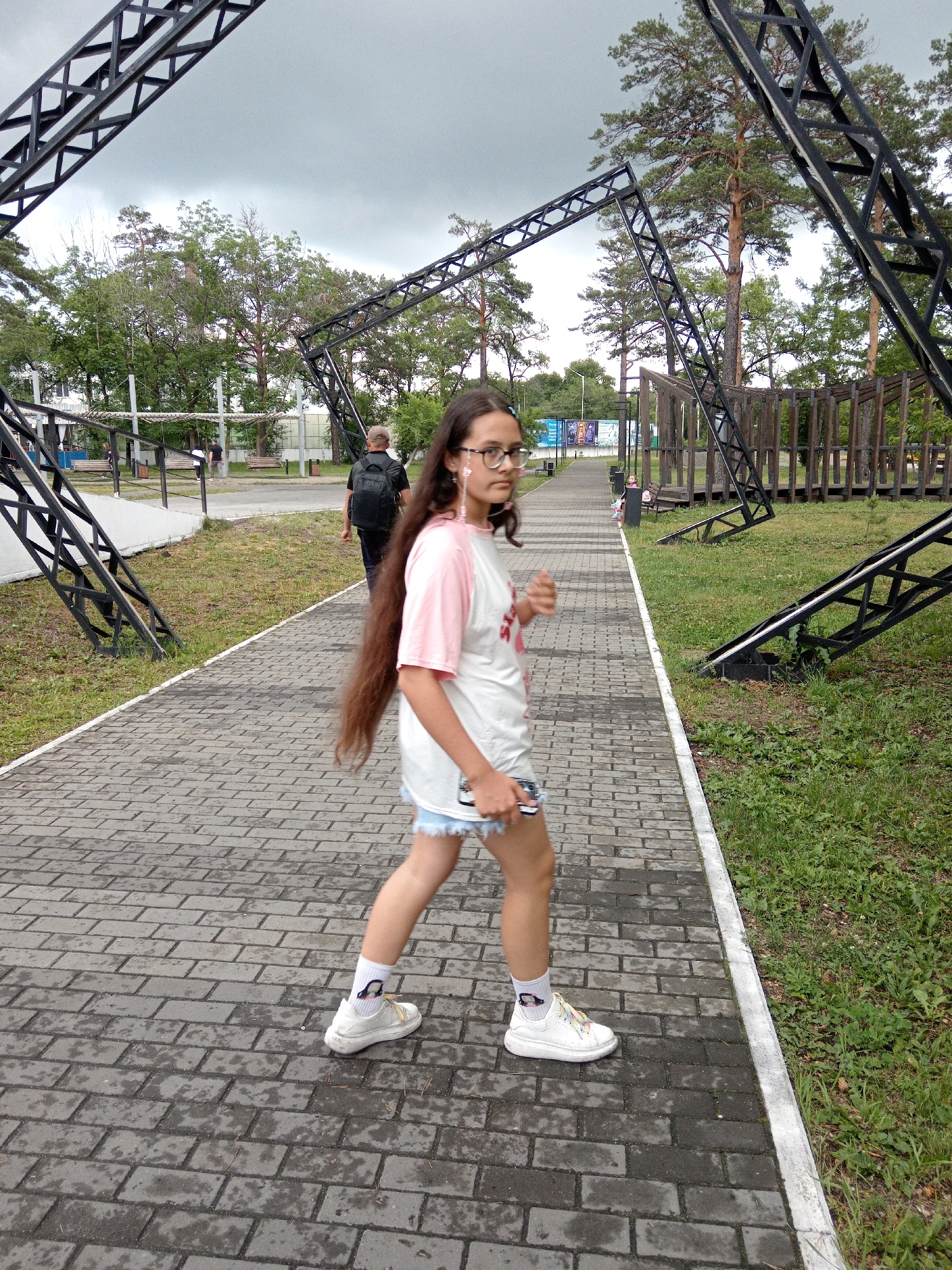 A young girl with long brown hair, wearing glasses, a pink and white t-shirt, and denim shorts, walks on a wet paved pathway. She is wearing white sneakers with colorful laces and white socks adorned with cartoon characters. She is also wearing earbuds with pink streamers. Behind her, a man with a backpack walks away from the camera. The pathway is bordered by green grass and tall trees. In the background, there are abstract metal structures and a wooden gazebo-like structure. The sky is overcast. The location appears to be a park or recreational area in the city of Belogorsk, Russia.