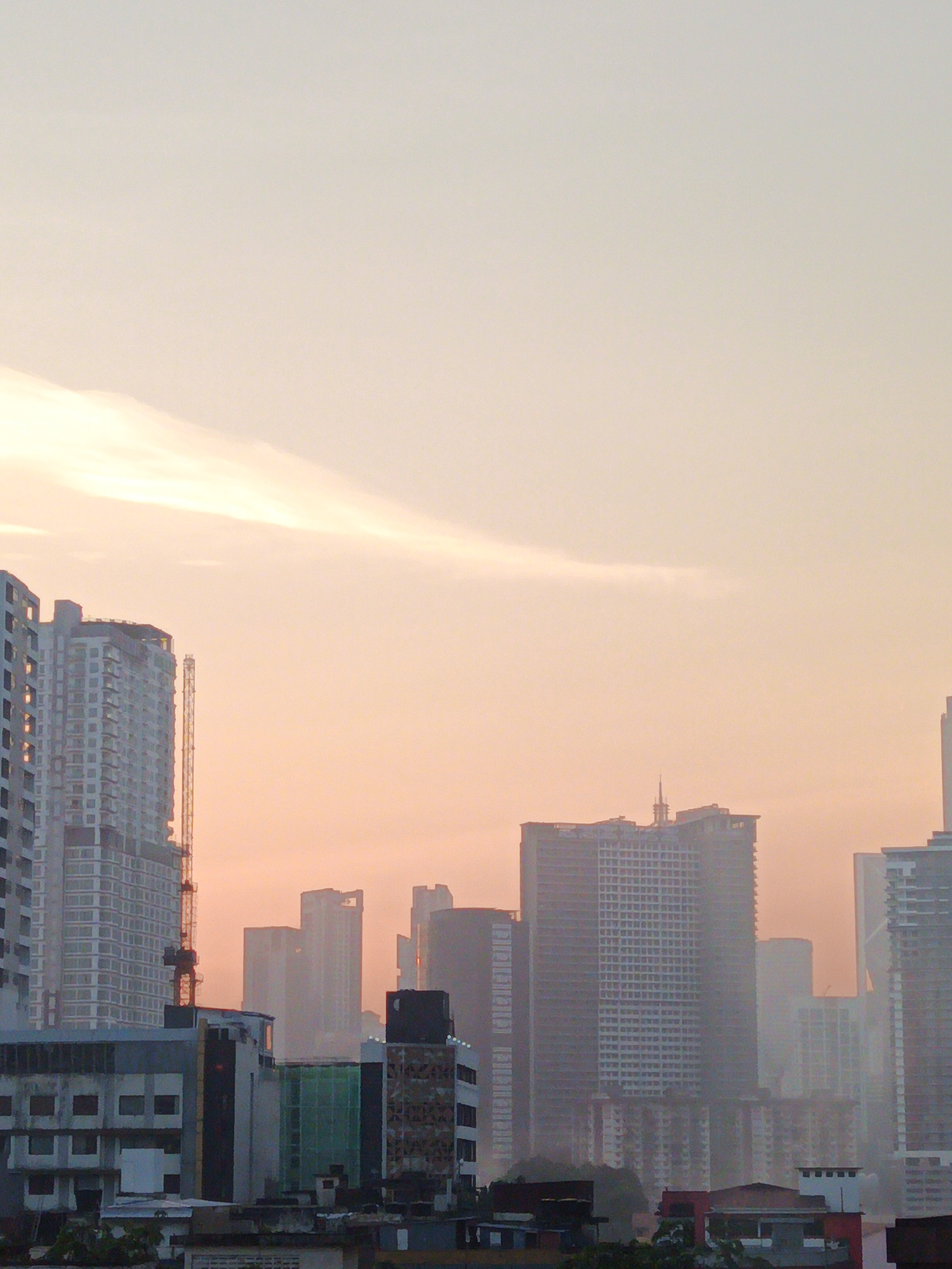 This is a vertical, medium-shot photograph of a cityscape at dawn or dusk in Kuala Lumpur, Malaysia. The sky is a gradient of pale yellow and soft orange, with a long, wispy cloud formation stretching horizontally across the upper half of the frame.
Dominating the lower portion of the image is a dense collection of modern high-rise buildings. On the left, a tall, light-colored apartment building with numerous rectangular windows is partially visible. To its right, another similar, albeit slightly shorter, building stands, featuring a prominent yellow construction crane extending upwards.
Further to the right, a cluster of skyscrapers of varying heights and architectural styles fills the middle ground. Some are sleek and modern with glass facades, while others appear more textured and complex. Several buildings appear to be under construction, indicated by their unfinished facades and exposed structural elements. Haze or light fog obscures the details of some of the buildings in the distance, creating a sense of depth.
In the foreground, a lower-rise section of the city is visible, with flat-roofed buildings, some with visible rooftop units or HVAC systems. The overall impression is one of urban development and the ongoing growth of the city, bathed in the warm, soft light of a sunrise or sunset. There are no people or animals visible in the image. The atmosphere is still and quiet.
