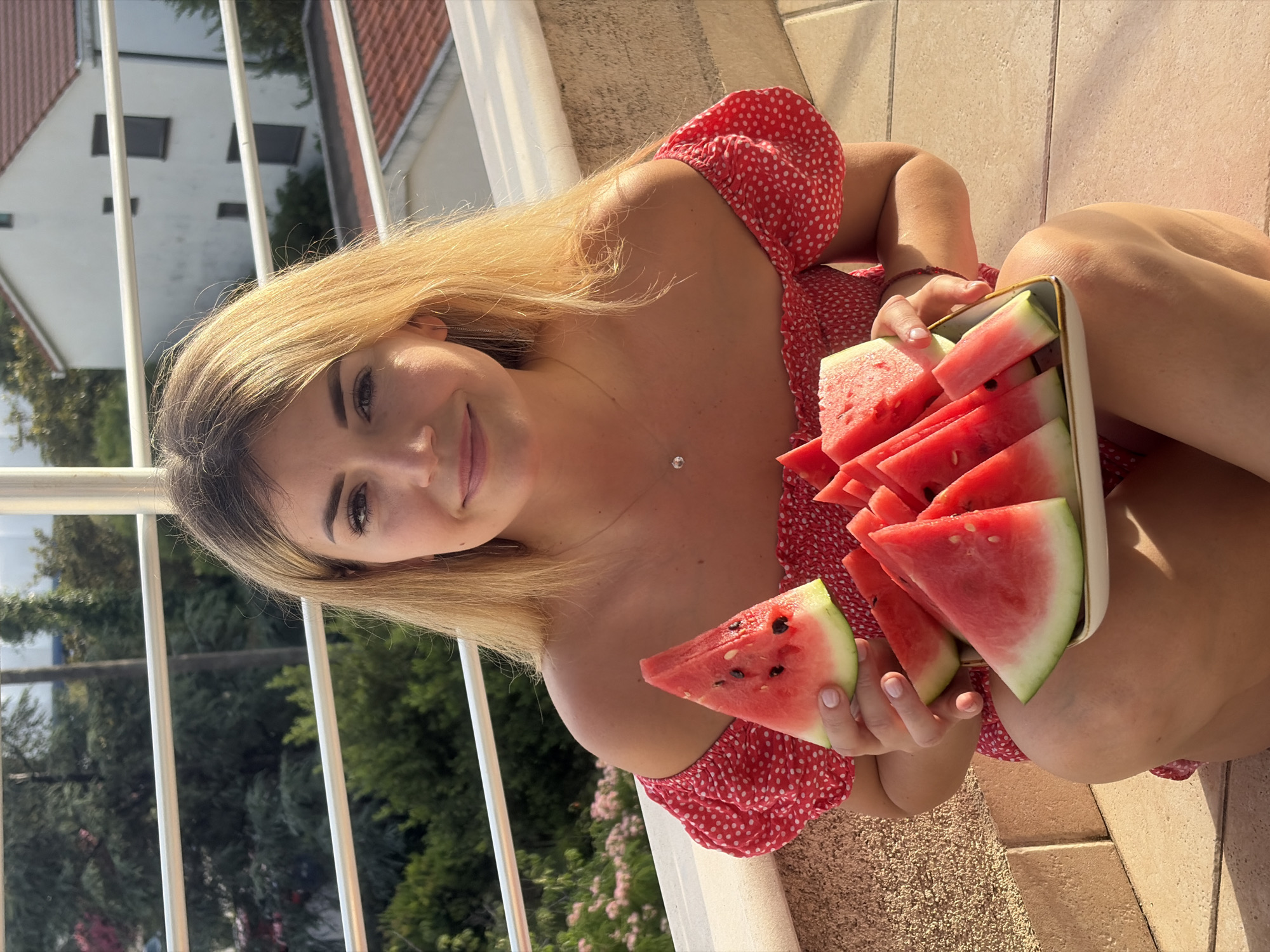 A young woman with long, blonde hair and a radiant smile is captured in a medium shot, looking directly at the camera. She is wearing a red off-the-shoulder top with white polka dots. Her left hand holds a triangular slice of watermelon, while her right hand supports a light-colored rectangular dish filled with numerous cut slices of watermelon. The watermelon slices are vibrant red with dark seeds and a light green rind.
The setting appears to be a sunny outdoor space, possibly a balcony or terrace, with a light-colored tiled floor visible in the foreground and to her right. A white railing runs vertically in the background, suggesting an elevated viewpoint. Beyond the railing, glimpses of a residential area are visible, including a white building with brown rooftops and dark rectangular windows, and lush green trees and foliage. Pink flowering bushes are also discernible in the distance. The scene is well-lit, indicating it is daytime, likely during late morning or afternoon given the warm, bright sunlight. The woman's expression is pleasant and relaxed, suggesting enjoyment of the moment and the refreshing fruit. There is no visible text in the image. The overall impression is one of a cheerful summer day in Budva, Montenegro.