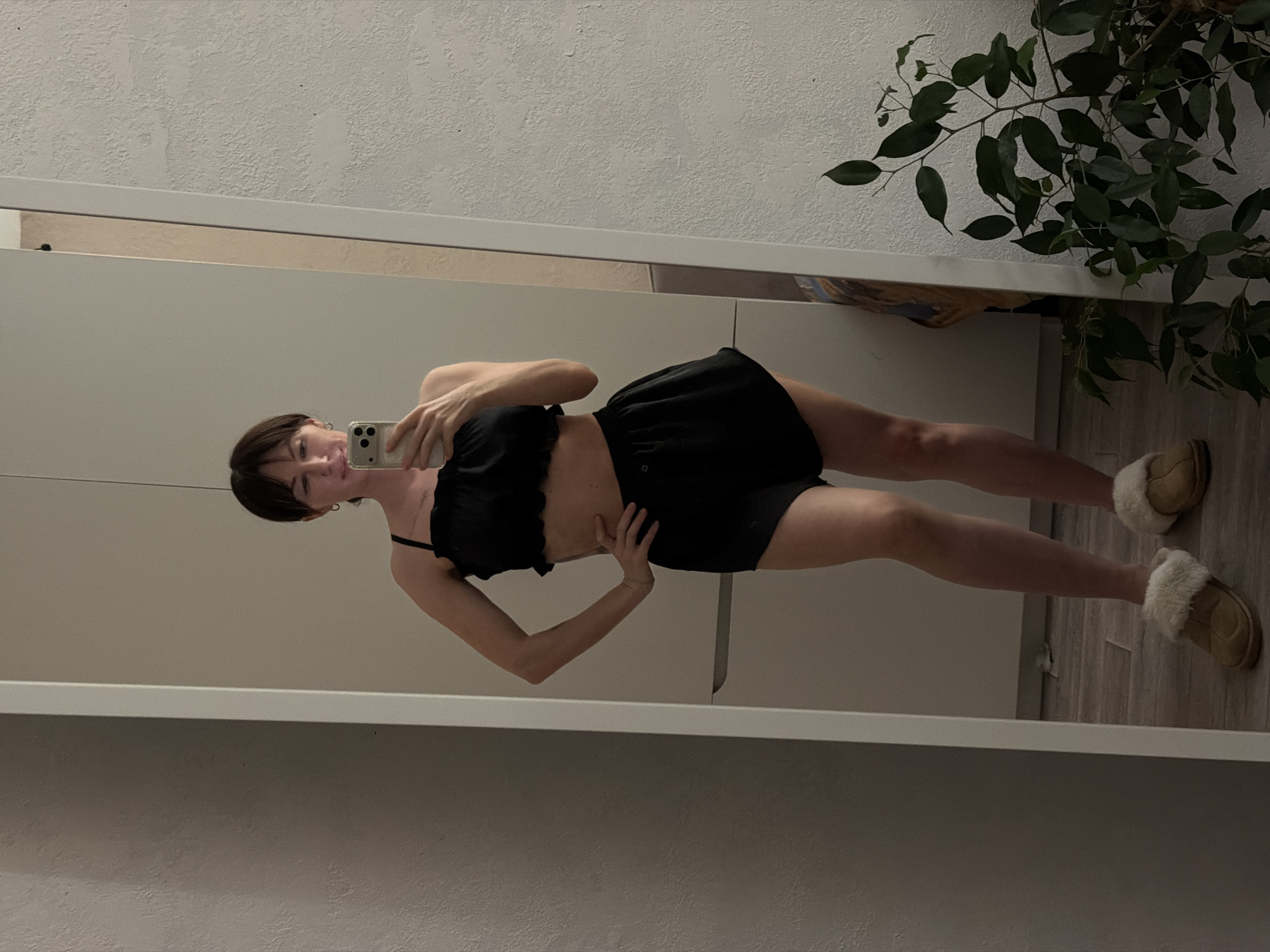 A woman is standing in front of a mirror, holding a smartphone and taking a selfie. She is wearing a black ruffled crop top and black shorts, and beige furry slippers. The background shows a wall with textured paint, a plant with dark green leaves, and a light-colored floor. The location is likely indoors, possibly in a home or apartment. The information about the city of Perm, Russia, is not evident in the image.