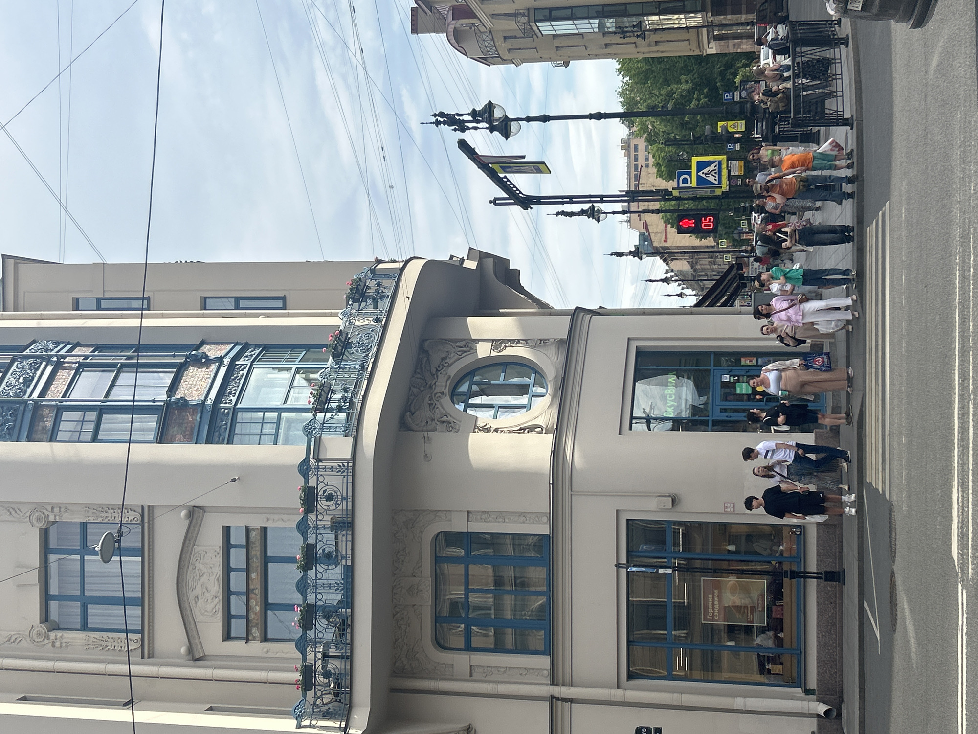 The image captures a bustling street scene at the intersection of Vosstaniya Street and Liteyny Avenue in Saint Petersburg, Russia, specifically focusing on the distinctive corner building at Vosstaniya Street 1 / Liteyny Avenue 60, known as the House of Fyodor Litvinov. The multi-story building, constructed in an Art Nouveau or eclectic style, features light-colored walls, numerous blue-framed windows, and decorative wrought-iron balconies adorned with pink potted flowers. A prominent curved bay window rises from the upper floors, while the ground floor displays an oval window on the corner facade and houses a "ВКУСВИЛЛ" (VkusVill) grocery store, along with another shop advertising "Горячие сэндвичи" (Hot Sandwiches).
In the foreground, an asphalt street with a white-marked pedestrian crosswalk is visible. A group of approximately 15-20 casually dressed pedestrians, including adults and a child, are gathered on both sidewalks. A smaller group stands near the VkusVill entrance, while a larger group waits at the right side of the crosswalk, observing a red pedestrian traffic signal displaying a "05" countdown. Overhead, a dense network of electrical wires crisscrosses the partly cloudy sky. Several ornate black streetlights are positioned along the street, some supporting traffic signals and blue "P" parking signs, along with yellow pedestrian crossing signs. On the far right, the rear of a dark-colored vehicle is partially visible. Buildings with a similar architectural style line the street in the background, interspersed with mature green trees.
