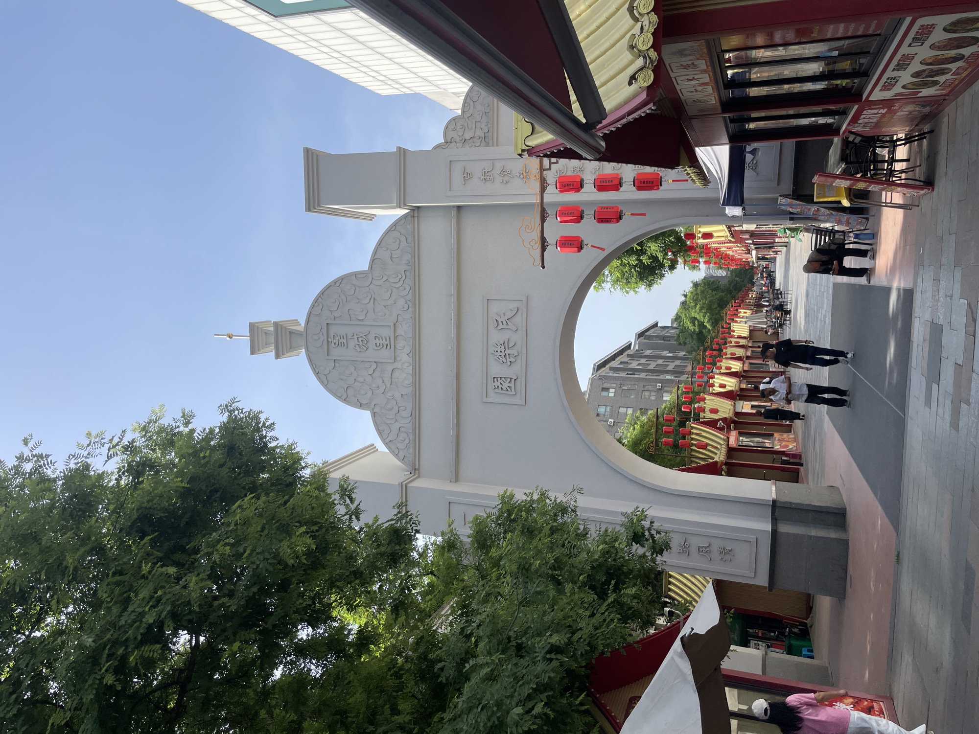 A low-angle shot looking down an urban street. An ornate, arched, white gate with Chinese characters and designs on its surface stands in the foreground. Red lanterns are suspended from the gate. The street is lined with shops and eateries, some with red awnings and signage. People are walking along the street. The sky is clear and blue. The scene appears to be in the Xincheng District of China.