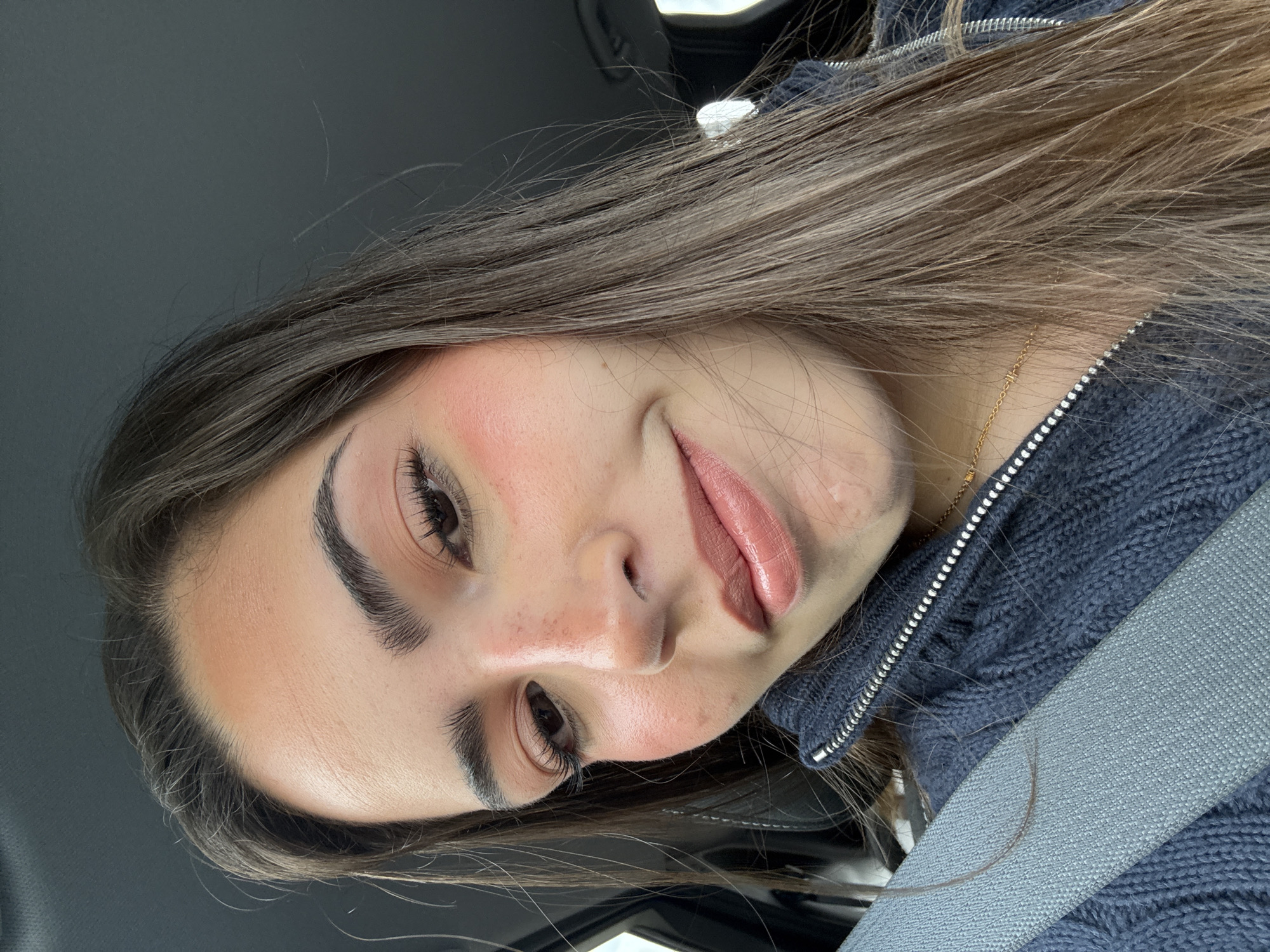 A young woman with brown hair and makeup is in a car, wearing a blue sweater and a seatbelt. The location is likely San Antonio, United States, based on the metadata.