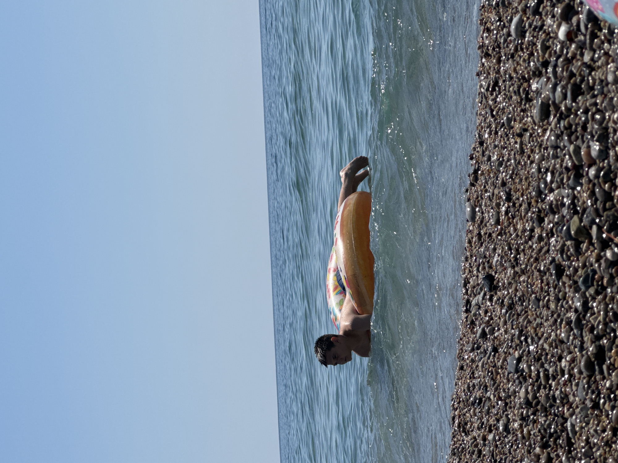 A young male with dark hair is floating on his stomach in the Adriatic Sea, supported by an inflatable ring. He is partially submerged in the water, with his head and shoulders visible. The water is a clear blue with small waves. In the foreground, there is a pebble beach. The sky is clear and blue. The location is the coast of Đurovići, Montenegro.