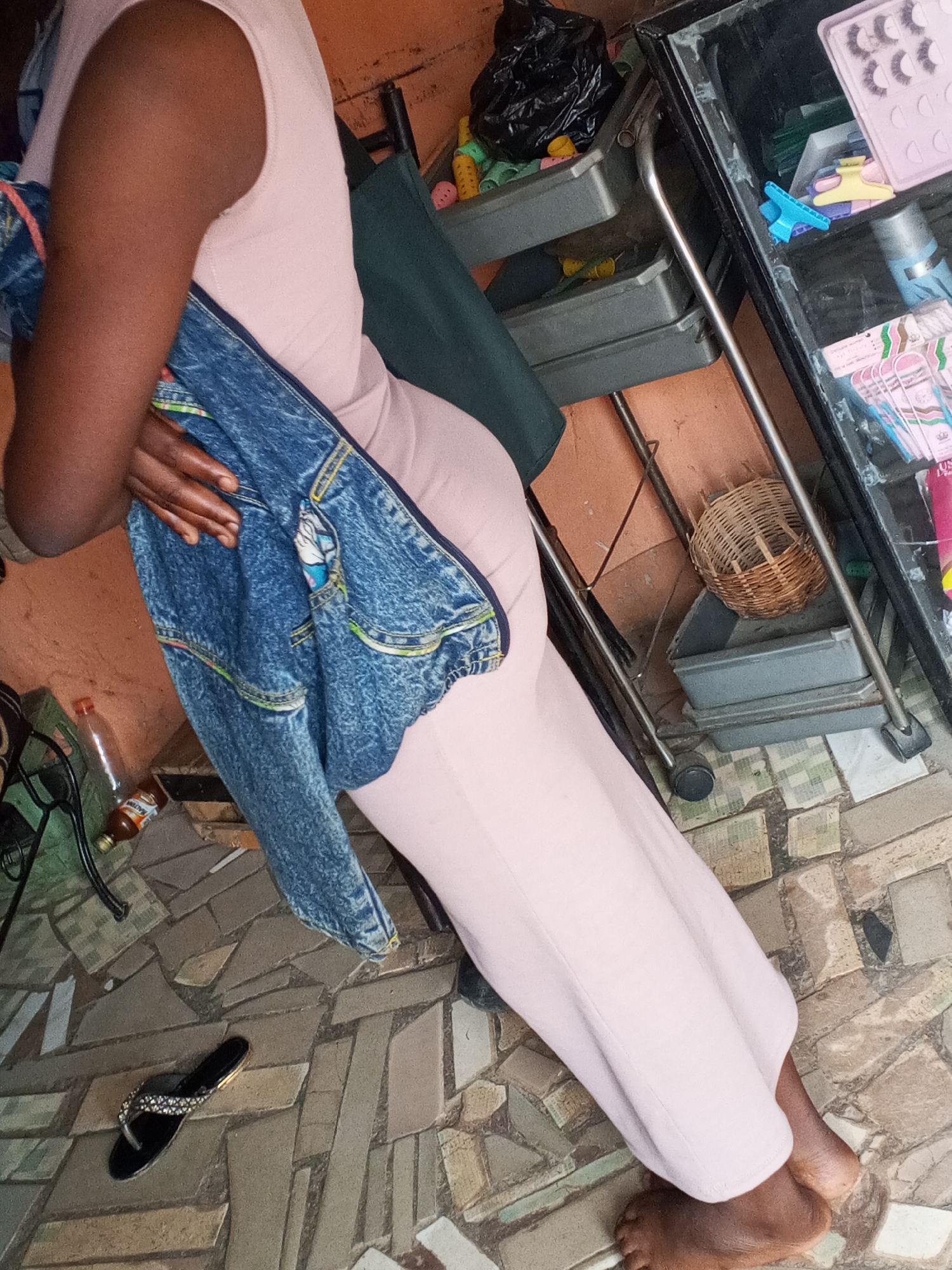 A woman with dark skin is wearing a light pink sleeveless jumpsuit. She is holding a blue denim jacket. To her right is a wheeled cart with trays containing various items including hair accessories and a black trash bag. The floor is tiled with mosaic tiles. There is a sandal on the floor. The location is Unknown, Unknown.