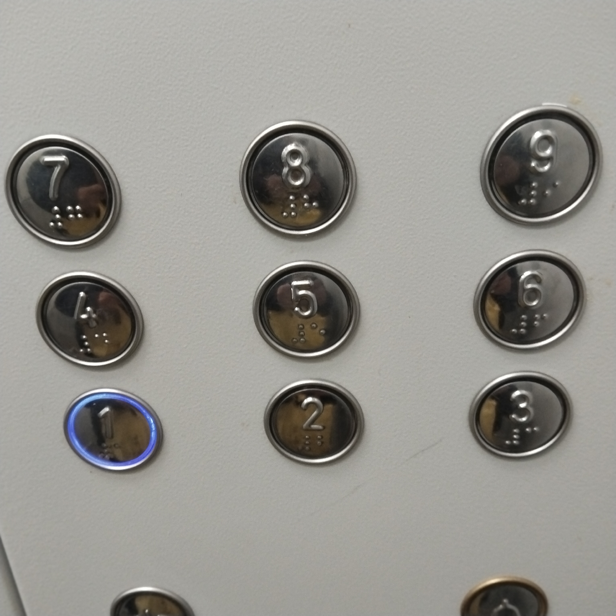 The image shows a close-up of an elevator panel with buttons numbered 1 through 9, arranged in a 3x3 grid. Button number 1 is illuminated with a blue light. Braille is present beneath each number. No people are visible. The background is a light-colored, possibly off-white, surface. There is no indication of the location in Chaikovsky, Russia.