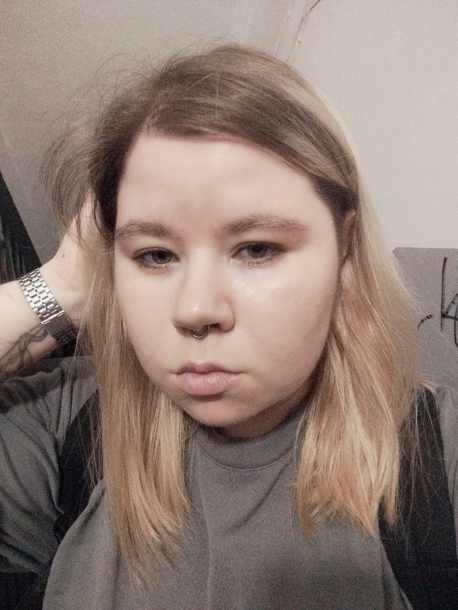 A close-up, indoor selfie of a person with light skin and blonde hair. The person wears a gray t-shirt, and their hair is slightly disheveled. They have a septum piercing and are wearing a silver wristwatch on their left wrist, which has a visible tattoo. The background consists of a white wall and a gray object, possibly a couch, with indistinct writing on it. The person's hand is raised, touching their head. The location is Unknown, Unknown.