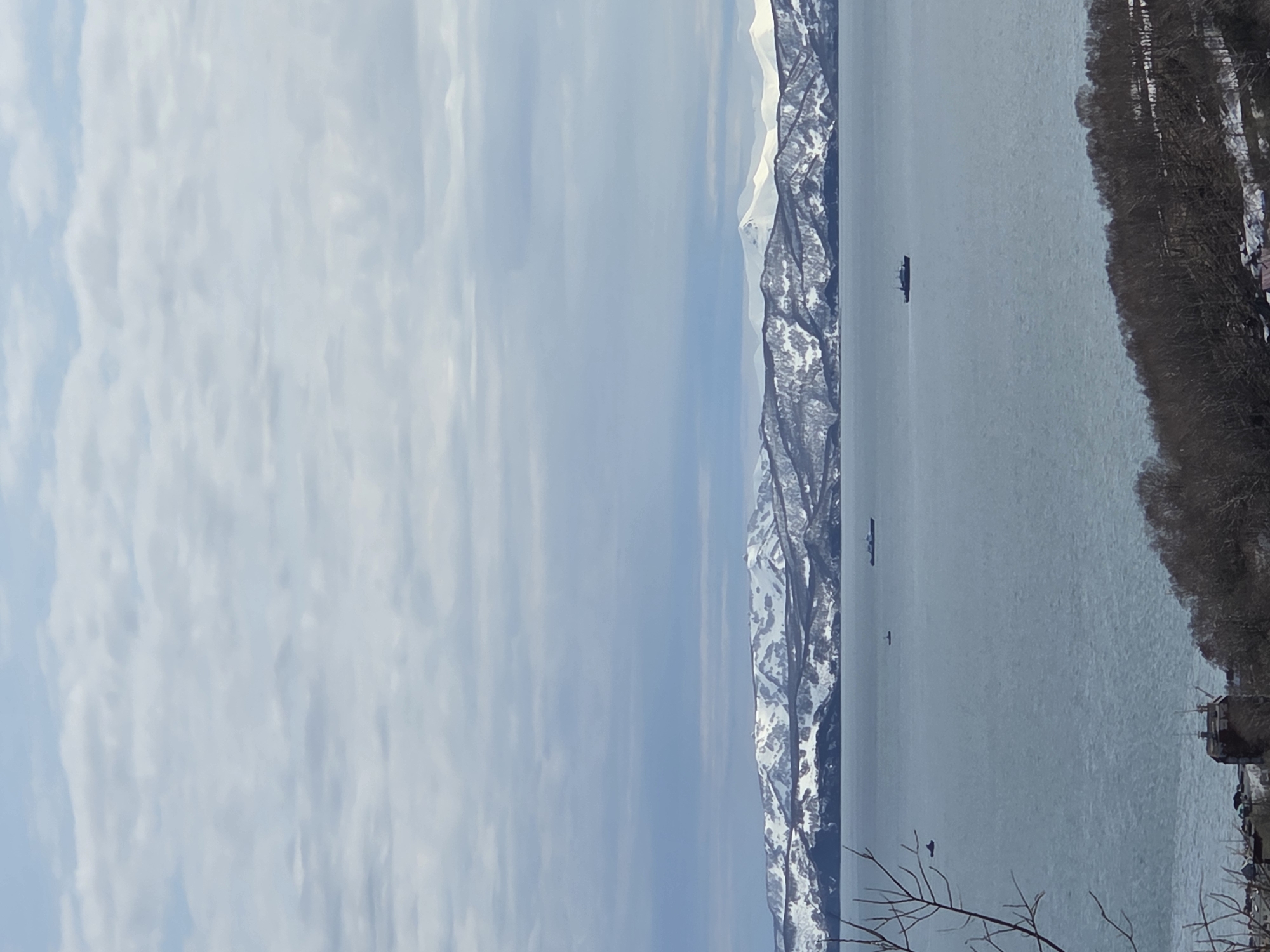 A serene landscape features a body of water, possibly a bay or sea, extending towards a rugged mountain range covered in snow. The sky is lightly clouded, and trees line the right side of the image. Several ships are visible on the water. The location is unidentified.